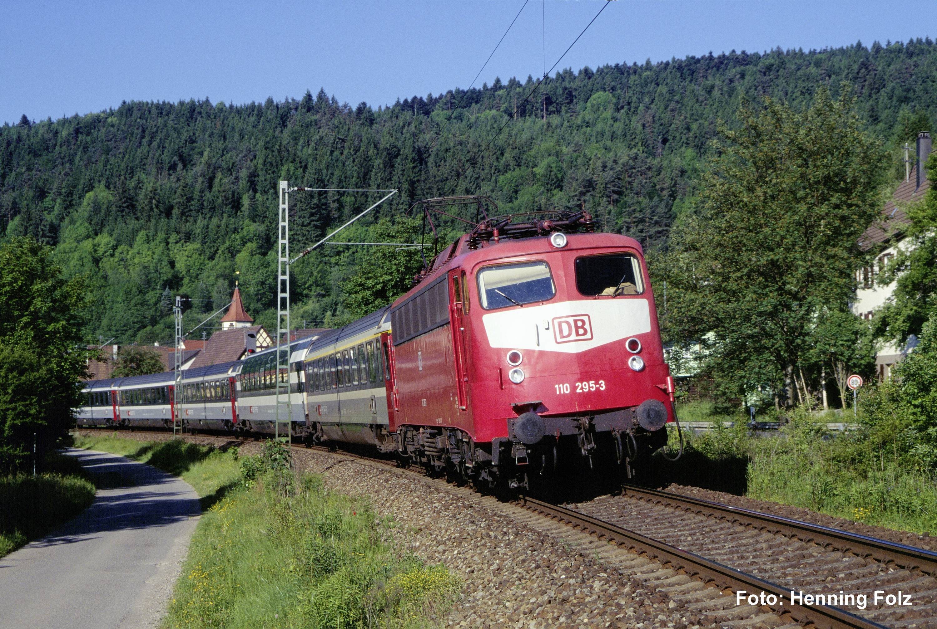 Un train de passagers DB rouge prend un virage sur des voies rurales à côté d'une route, avec des collines boisées et des maisons en arrière-plan.