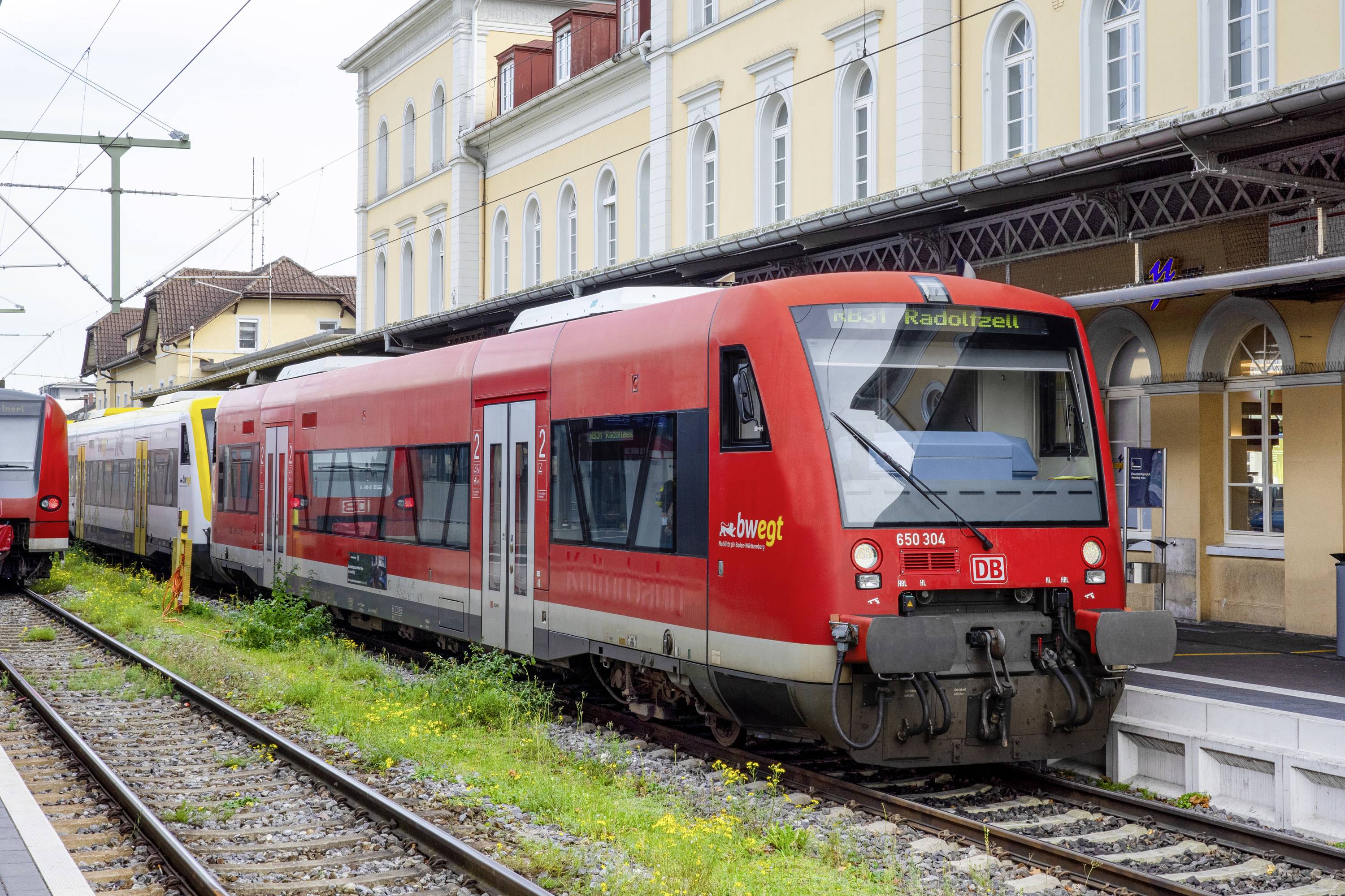 Train régional rouge sur un quai de gare avec des fils aériens et des bâtiments historiques en arrière-plan.