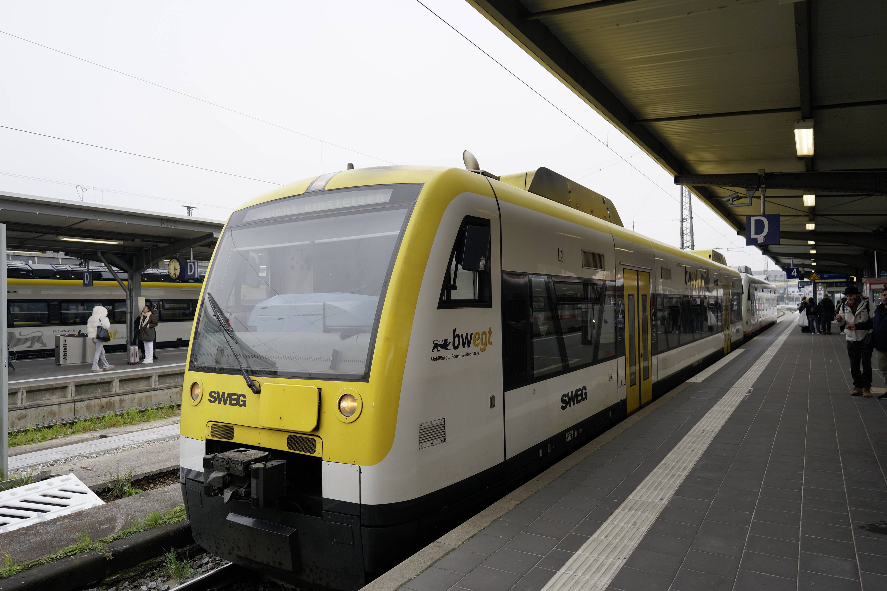 Train de passagers SWEG jaune et blanc à une gare, vue de l'avant-gauche avec des voyageurs attendant à proximité.