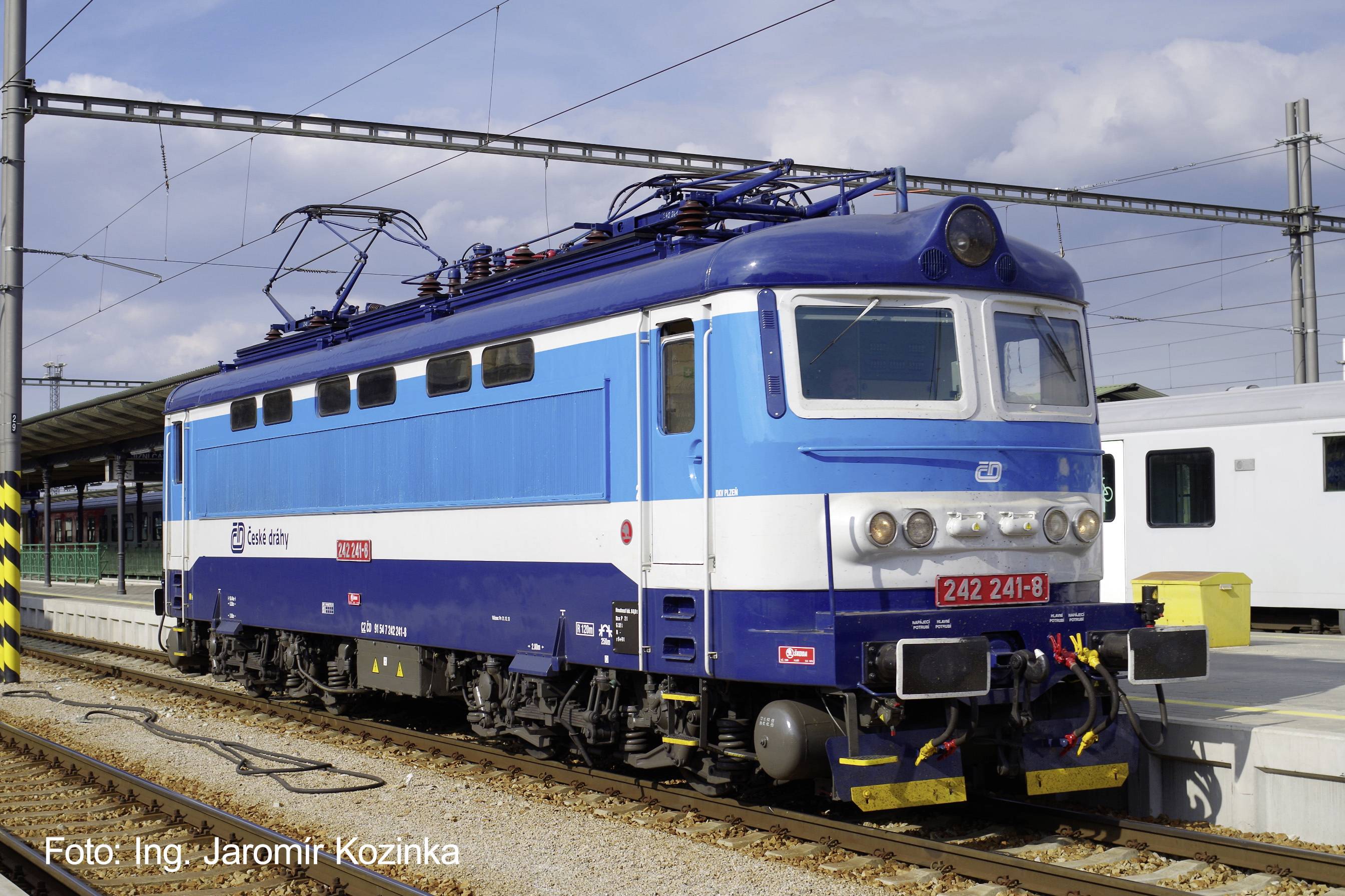 Locomotive électrique bleue garée sur un quai de gare ferroviaire, vue de face-gauche avec les lignes électriques aériennes visibles.