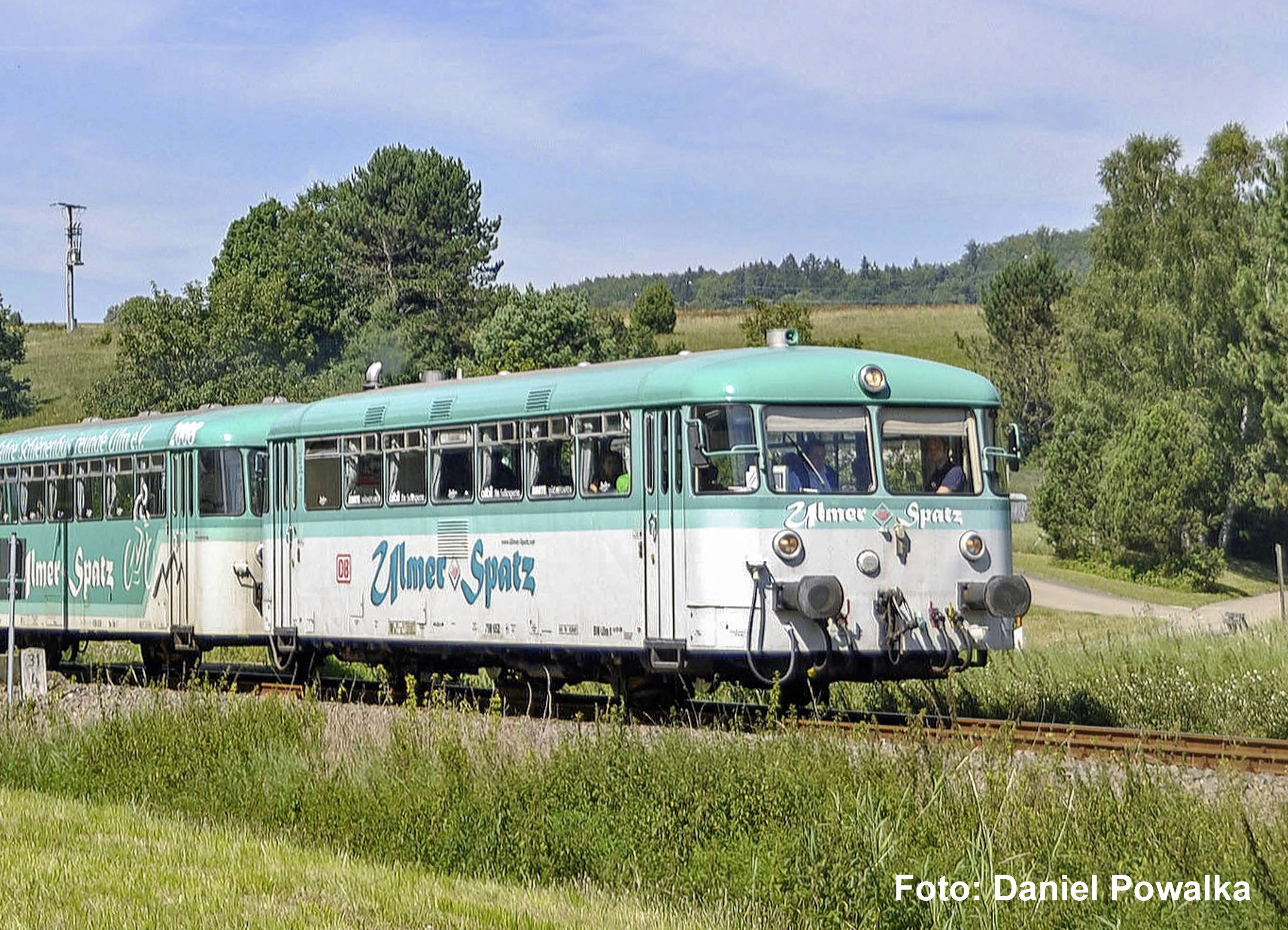 Un train de passagers bleu-vert et blanc étiqueté « Umer Spatz » voyage à travers un paysage rural verdoyant sous un ciel bleu.