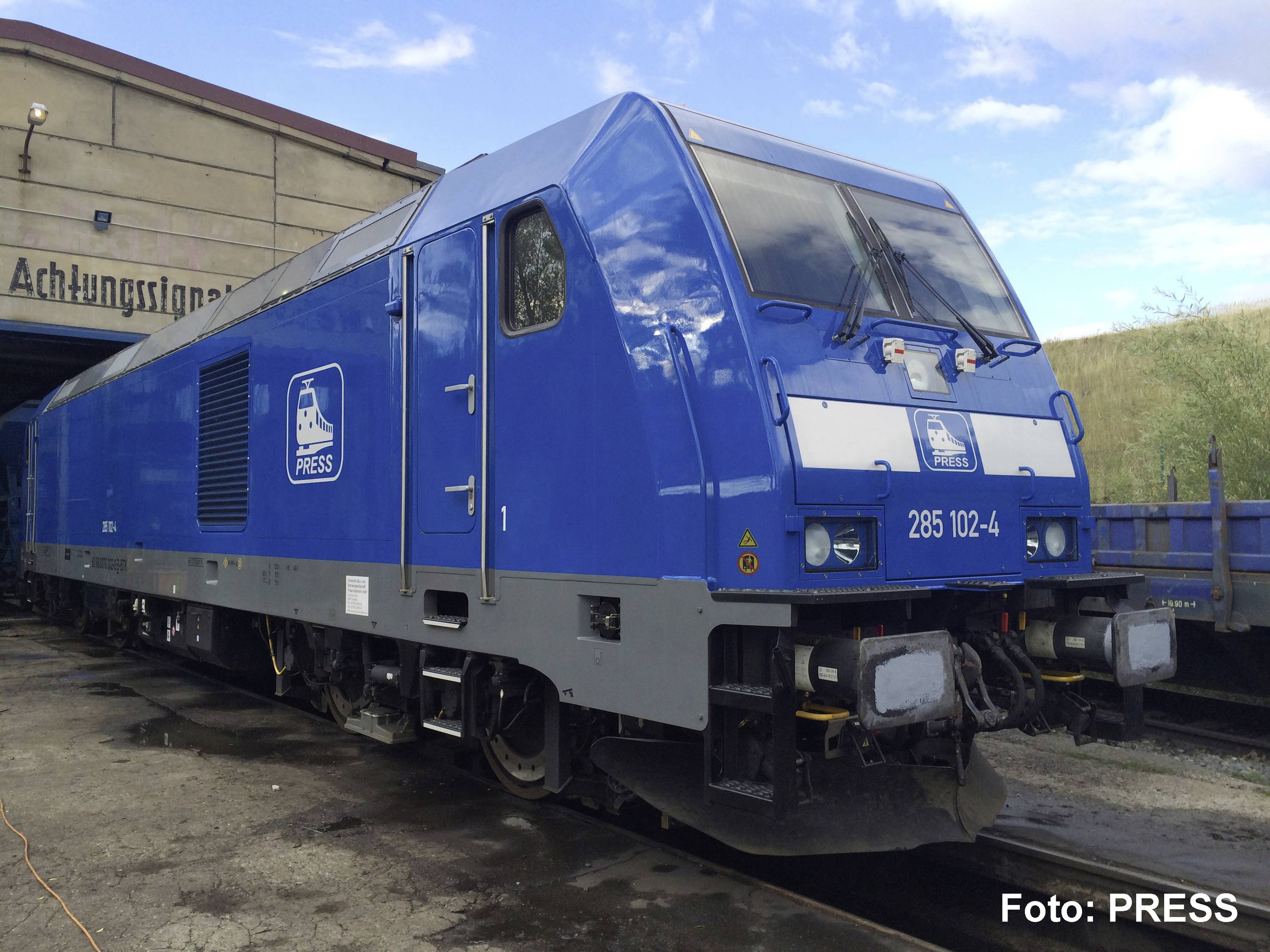 Locomotive de fret bleue avec des logos « PRESS » sur le côté, garée à l'extérieur d'un dépôt sous un ciel partiellement nuageux.
