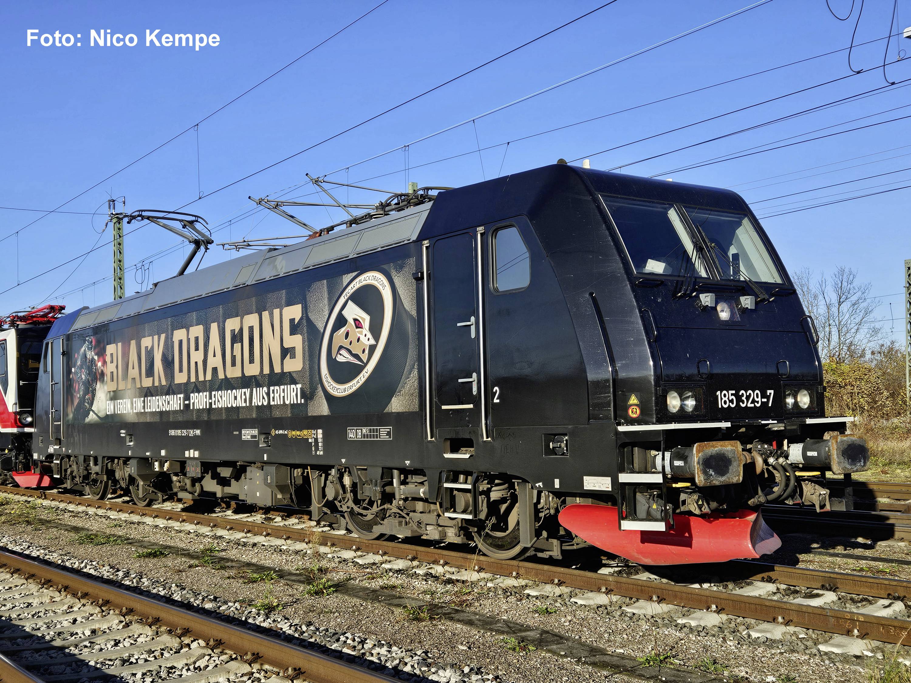 Locomotive électrique noire étiquetée « BLACK DRAGONS » sur les rails de chemin de fer sous des lignes aériennes d'alimentation électrique, avec un chasse-neige rouge et un grand logo sur le côté.