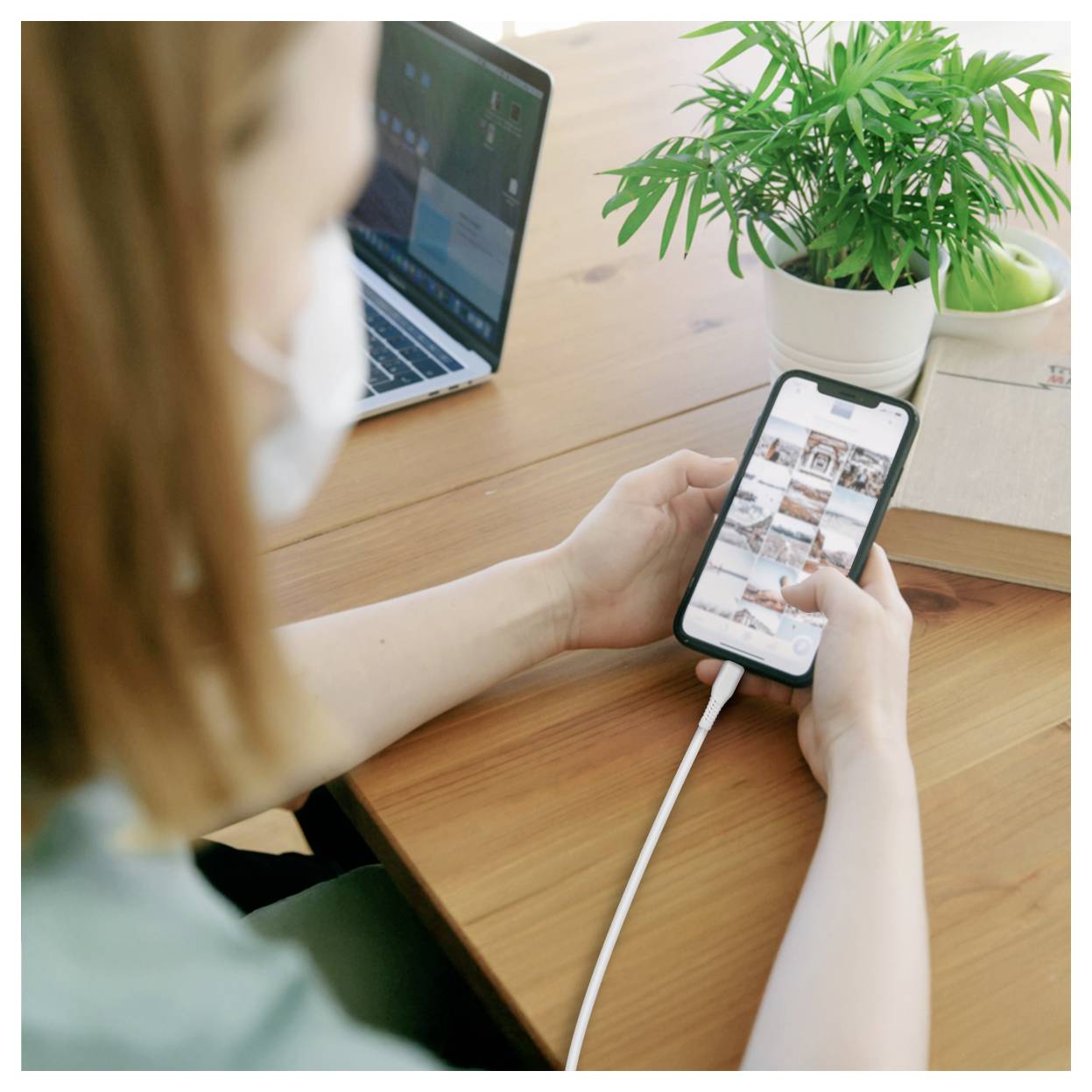 Une personne portant un masque consulte un flux de photos sur son téléphone à un bureau à côté d'un ordinateur portable et d'une plante en pot, le téléphone en train de se charger.