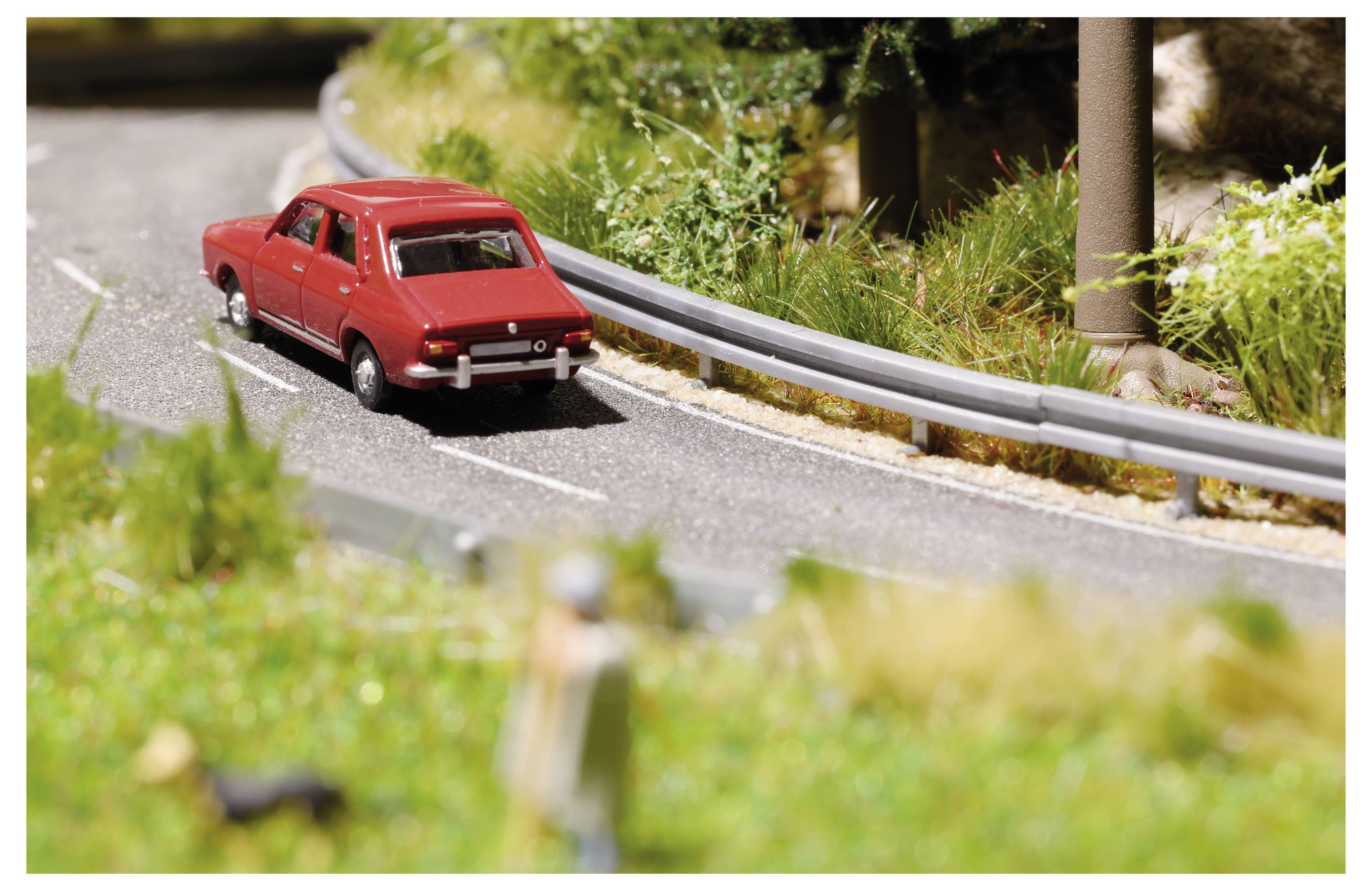 Voiture jouet rouge roulant le long d'une route miniature sinueuse à côté d'une barrière de sécurité, avec de l'herbe et des arbres flous en arrière-plan.