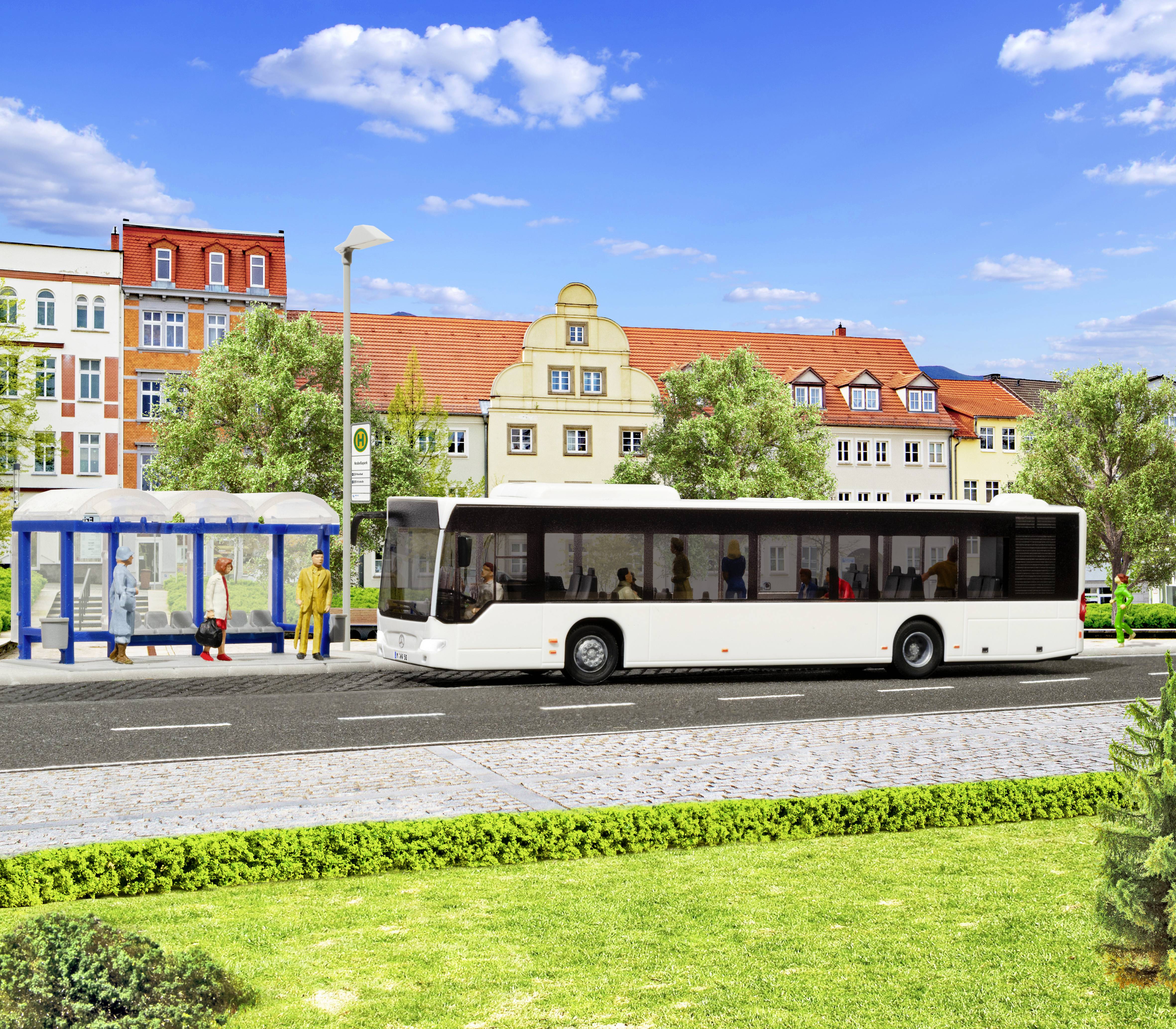 Un bus blanc s'est arrêté à côté d'un abri de bus couvert sur une rue bordée de bâtiments colorés et d'arbres par une belle journée ensoleillée.