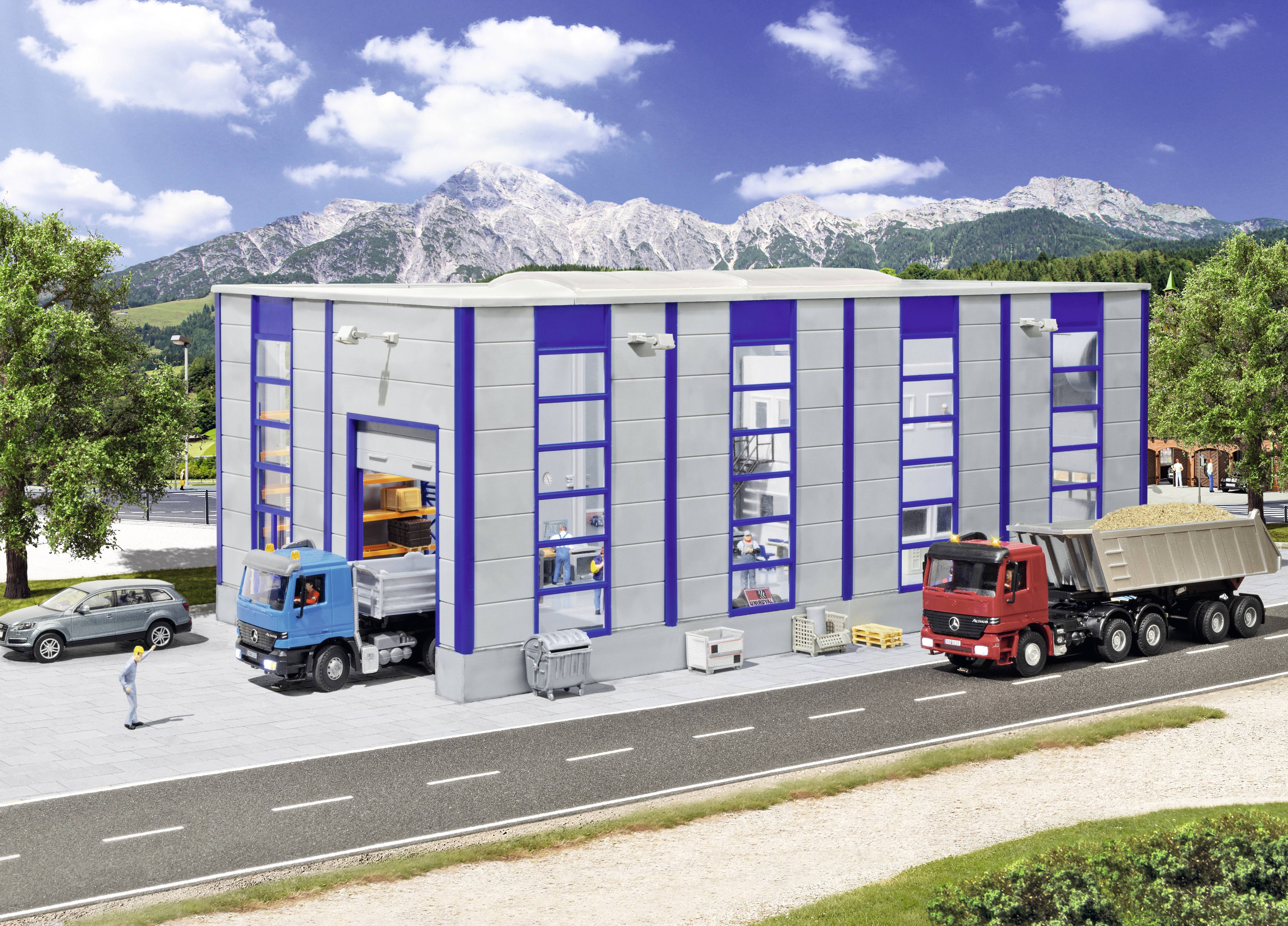 Entrepôt industriel avec quais de chargement ouverts, un camion bleu en cours de déchargement et un camion benne rouge garé à l'extérieur, situé face aux montagnes et un ciel bleu.