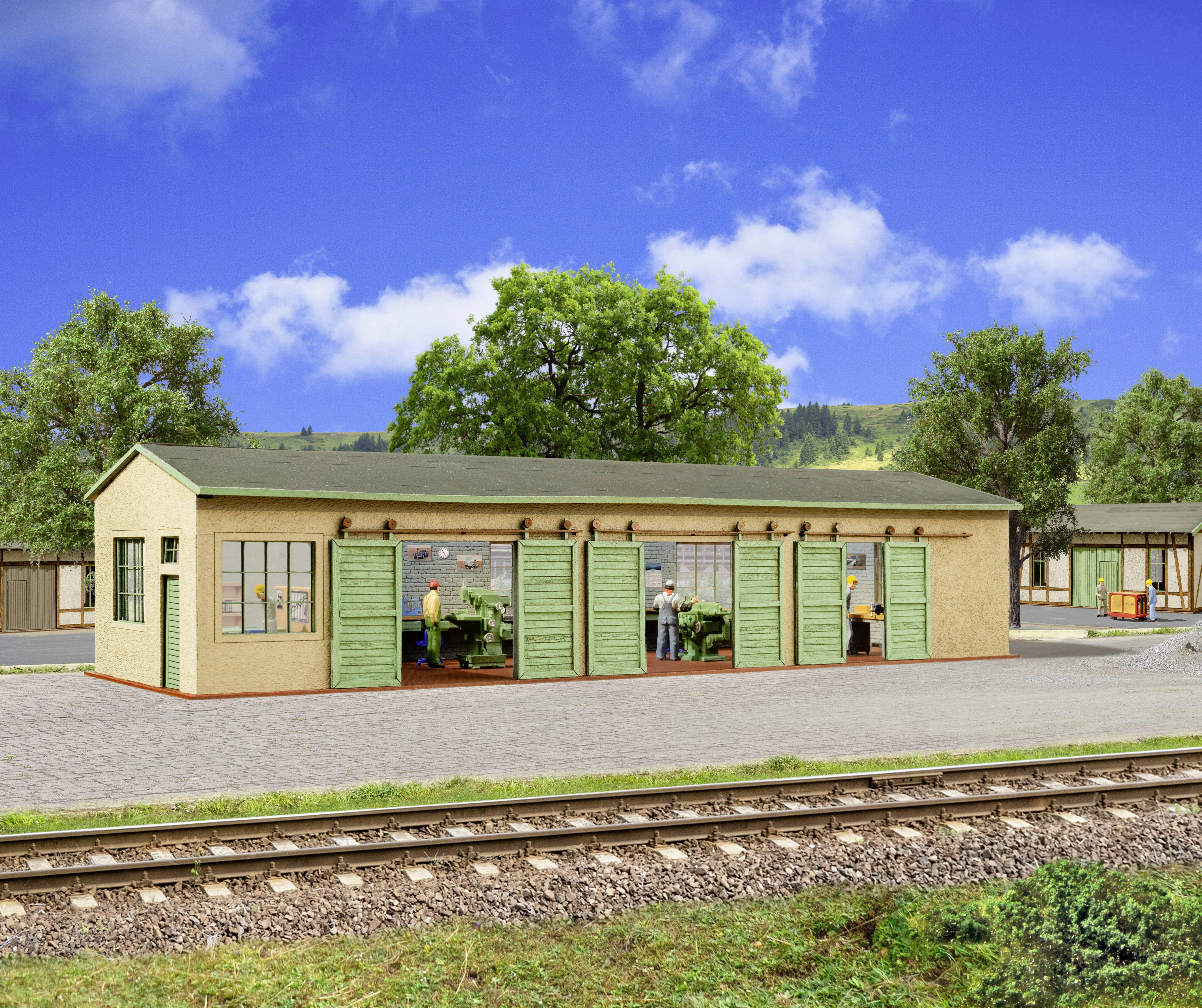 Quai de gare modèle avec un petit bâtiment beige et des portes vertes, des travailleurs à l'intérieur, des voies ferrées au premier plan, des arbres et un ciel bleu en arrière-plan.