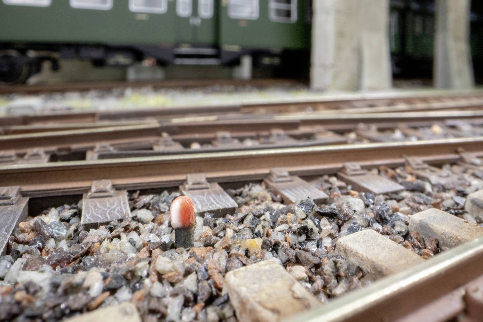 Gros plan des voies de chemin de fer miniature avec ballast de gravier et un petit marqueur de voie rouge et blanc ; wagons de train flous en arrière-plan.