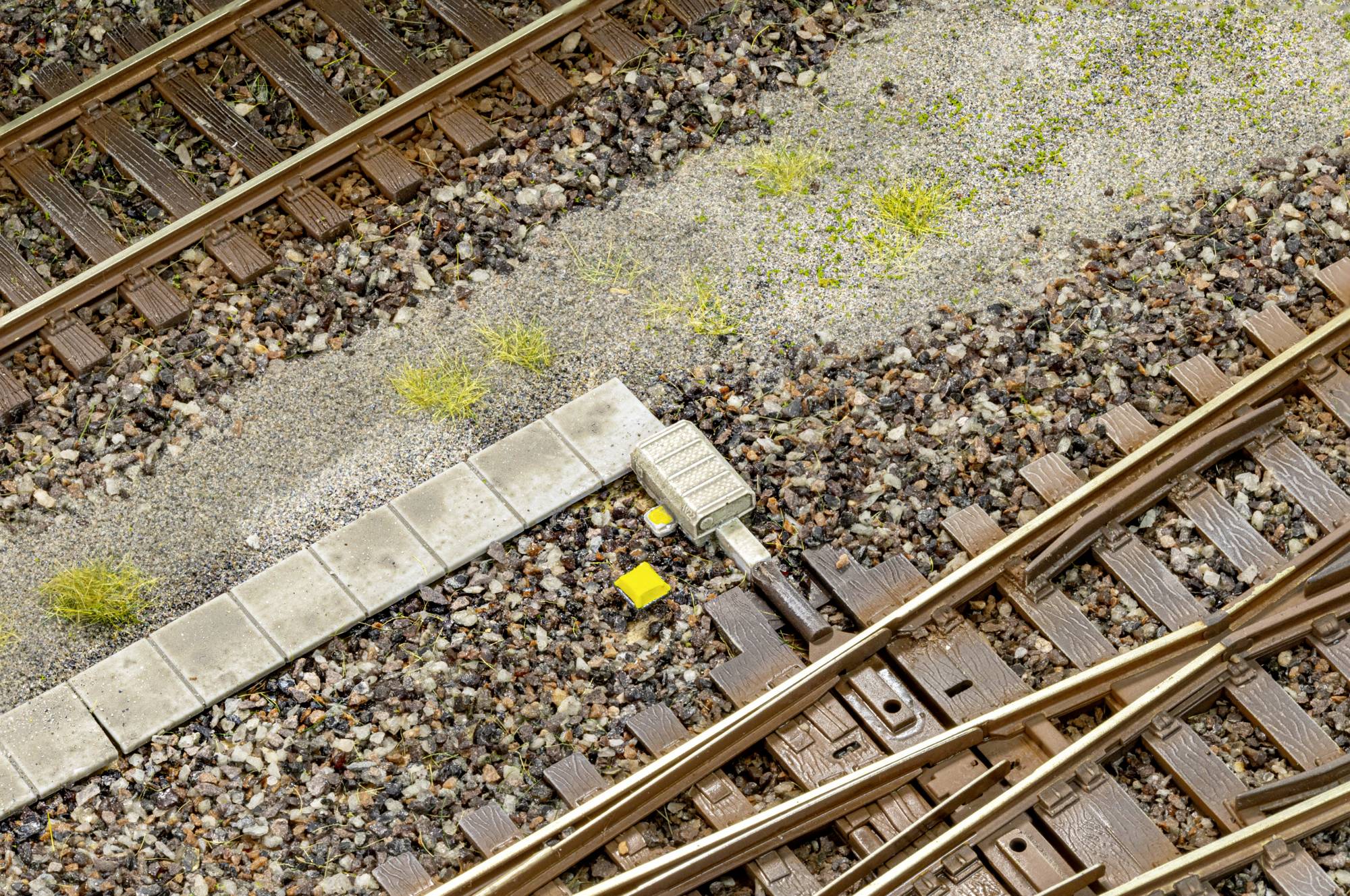 Croisement de rails de chemin de fer au niveau d'un aiguillage, avec ballast, bordure en béton et un petit marqueur de voie jaune près de la jonction.