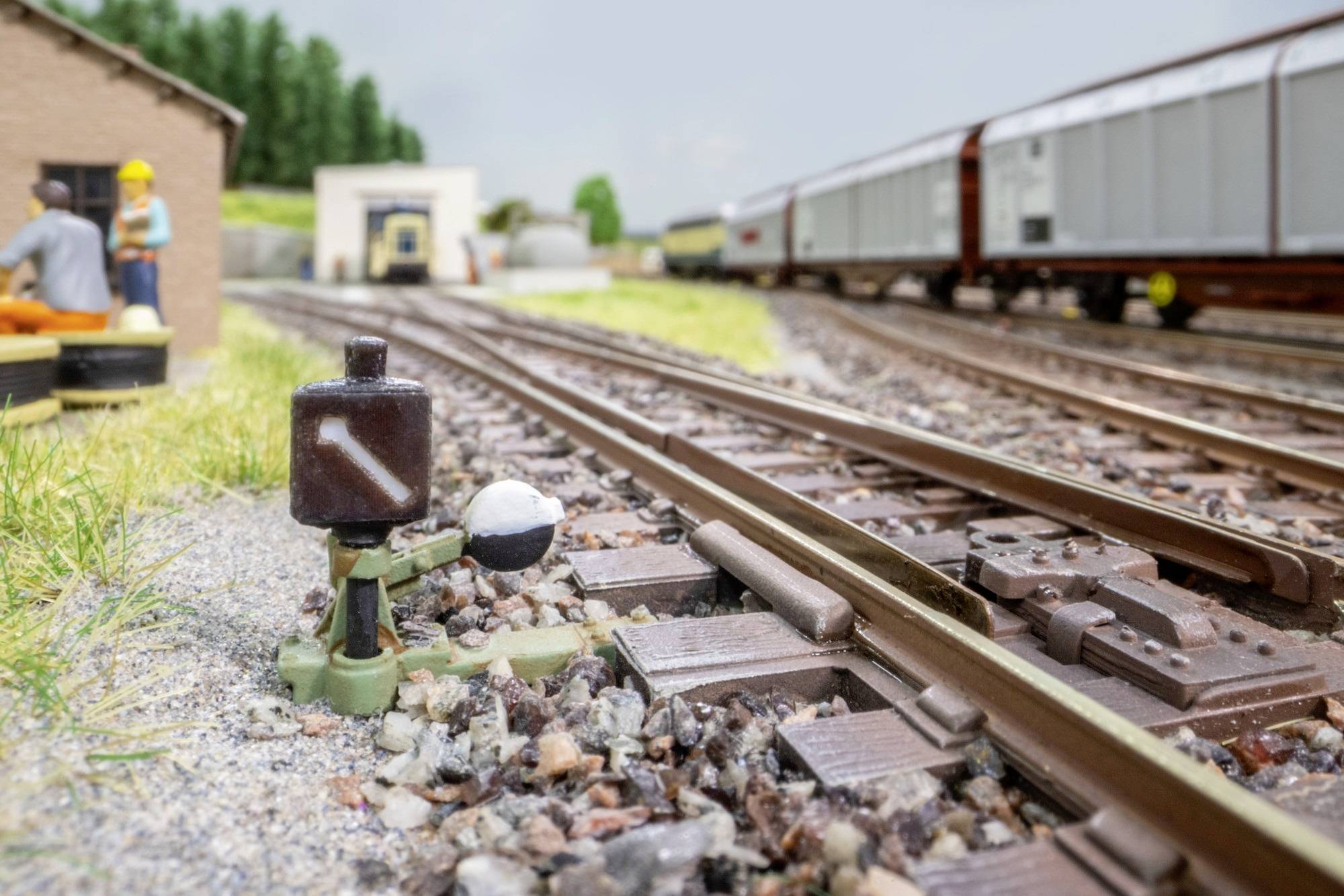 Voies de chemin de fer miniature avec un train de marchandises en arrière-plan et petites figurines d'ouvriers à côté d'un bâtiment, présentées dans une cour de triage ferroviaire miniature.