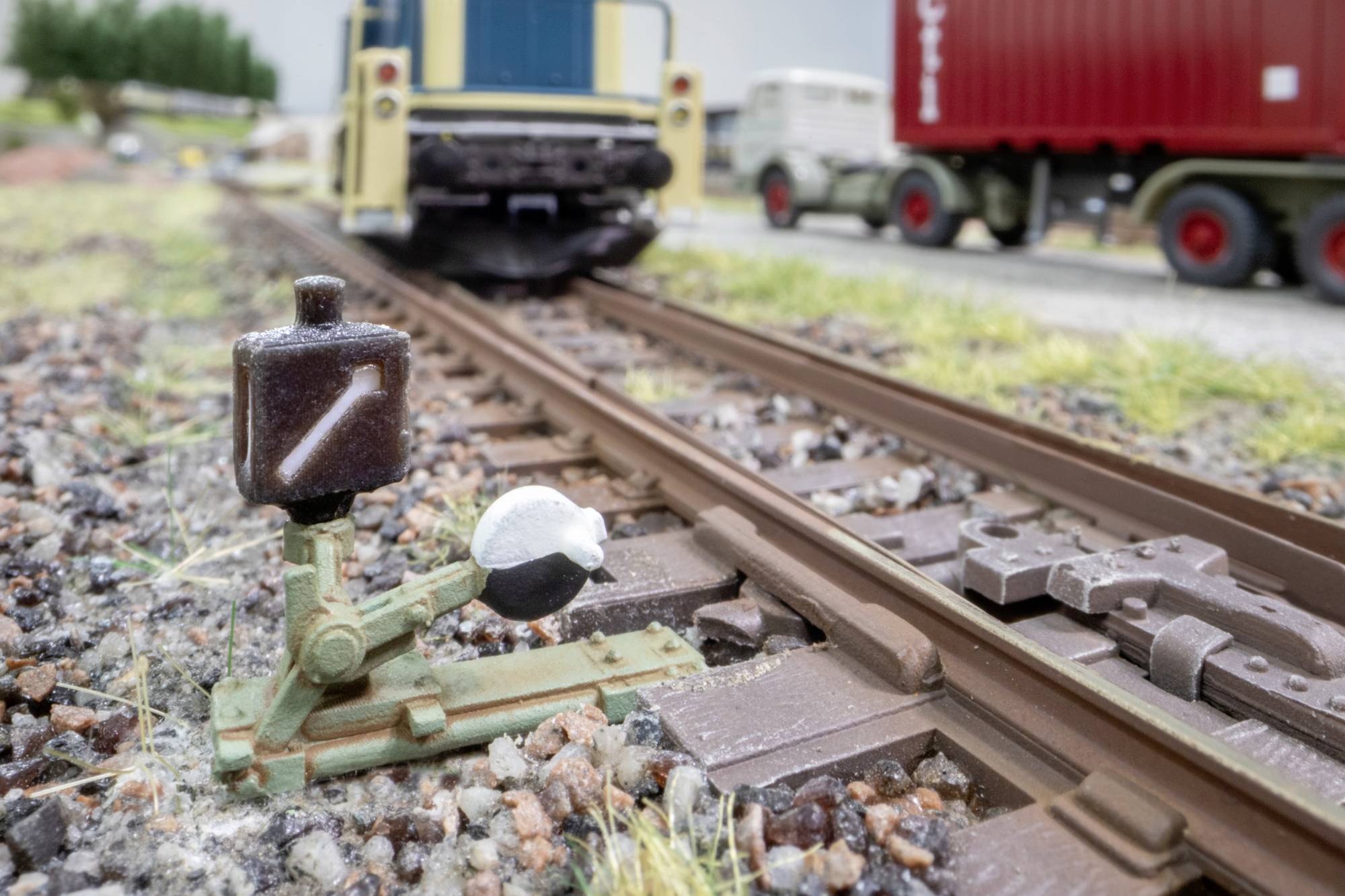 Gros plan d'un aiguillage de train miniature avec un levier de signal devant une locomotive jaune, un wagon-citerne blanc et un wagon de marchandises rouge sur des voies parallèles.
