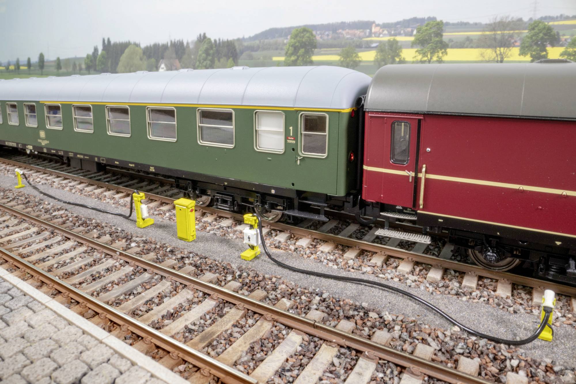 Deux voitures de train de passagers de modèle réduit couplées, verte et rouge, sur des voies d'exposition avec des câbles électriques visibles et de petites cabines de signalisation.