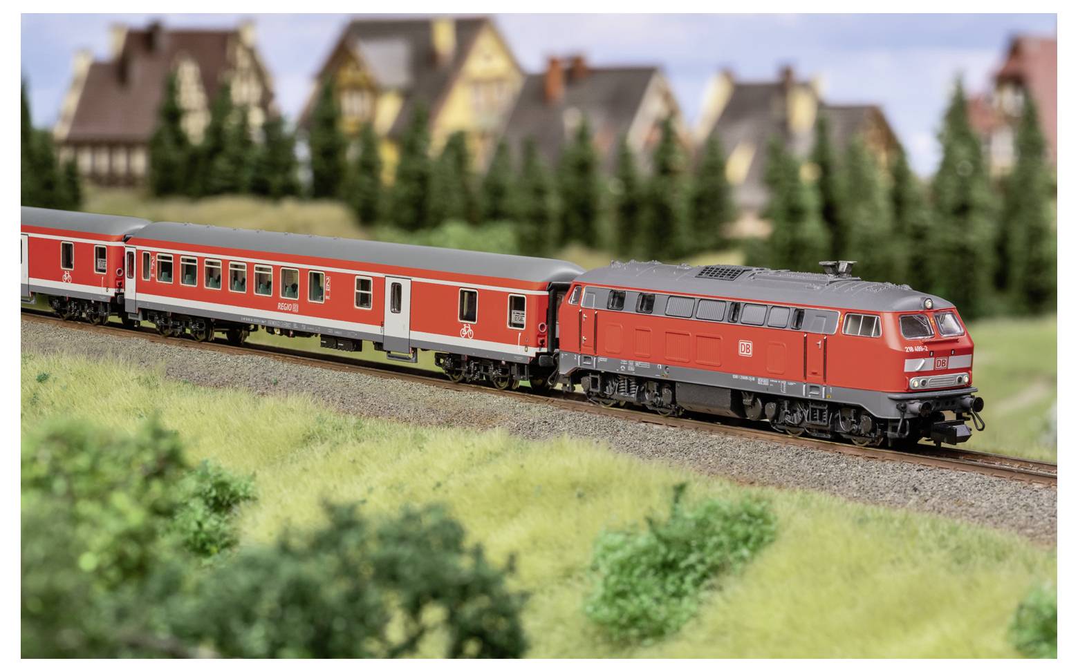 Train miniature rouge tirant des voitures de passagers rouges le long d'une voie courbe à côté d'un paysage herbeux, avec des maisons de village floues en arrière-plan.