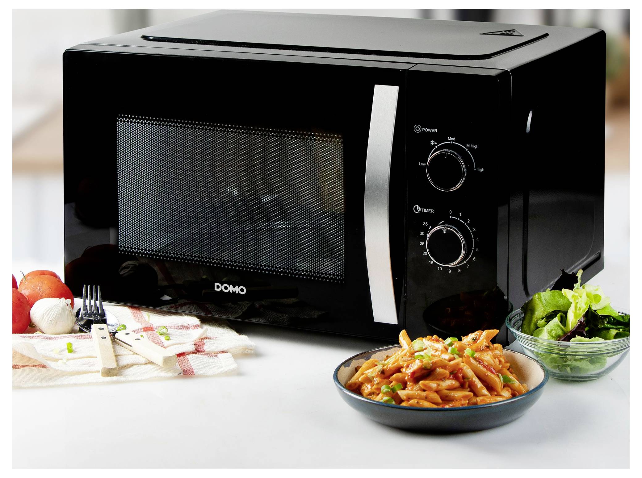 Black countertop microwave oven with two control dials, shown with a bowl of pasta, salad, and a place setting on a white table.