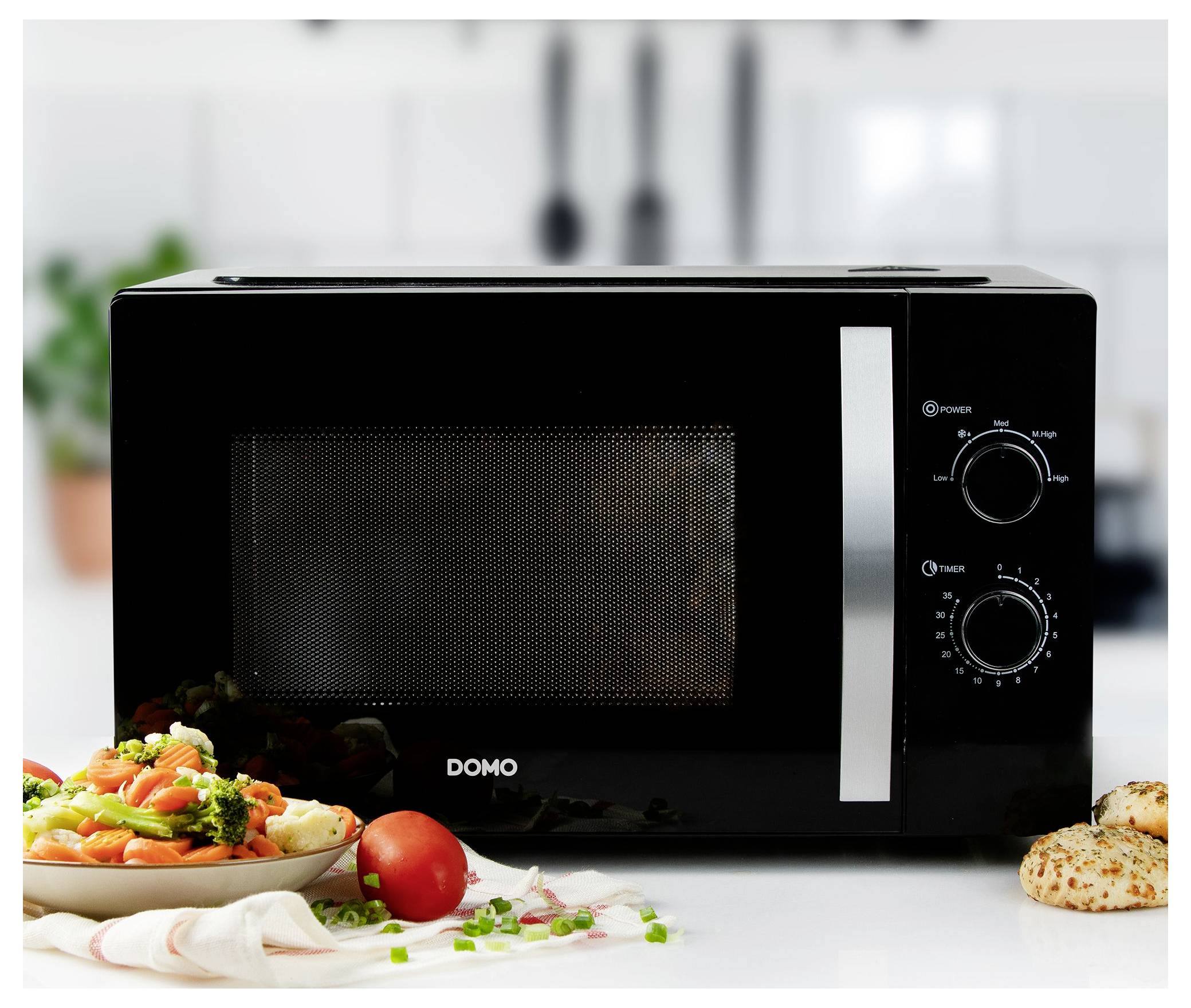 Black microwave oven with two control knobs on a kitchen counter, surrounded by plates of prepared food and vegetables.