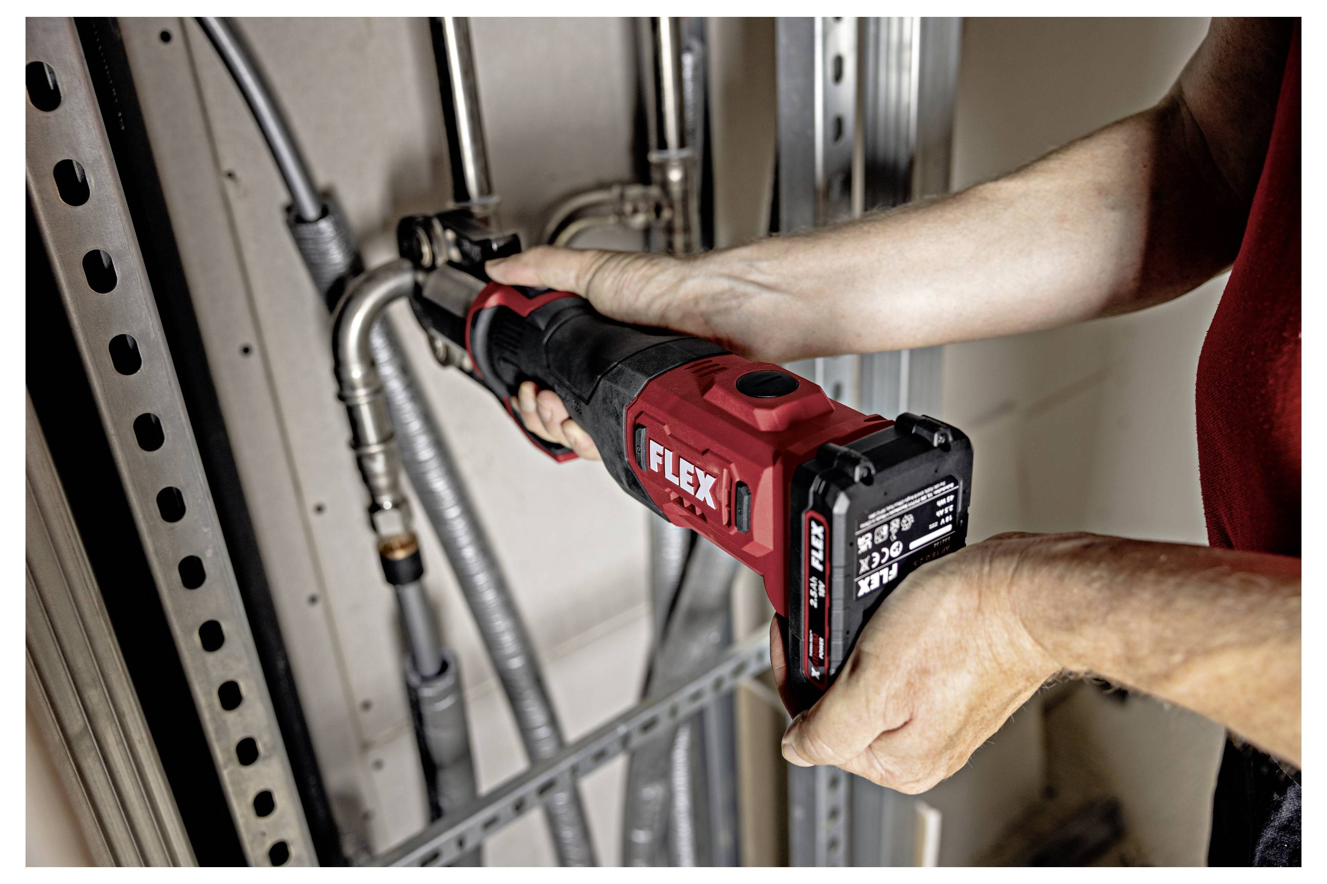 Personne utilisant une scie réciproquante sans fil FLEX rouge pour couper un conduit métallique à l'intérieur d'un mur ou d'un cadre utilitaire.