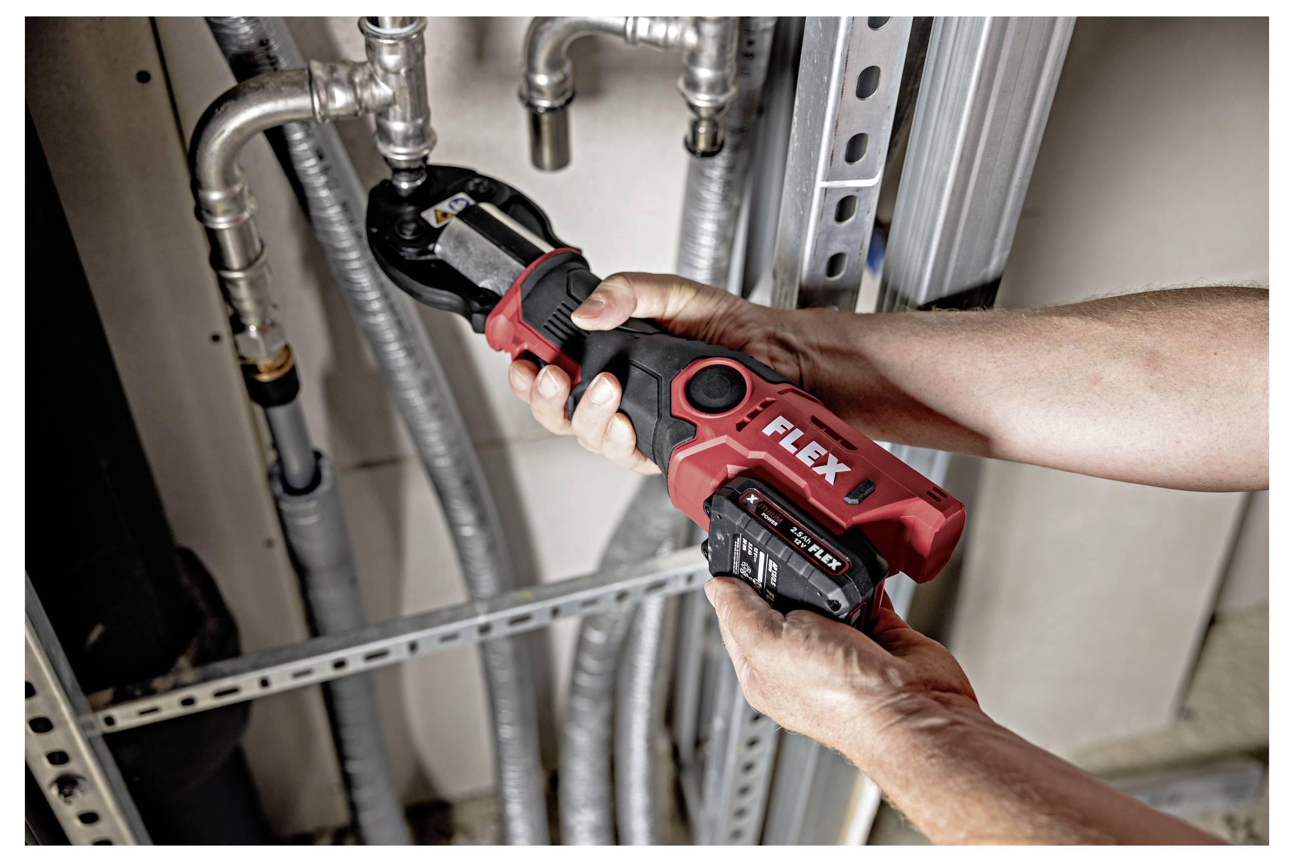 Des mains utilisant une scie à tuyau sans fil FLEX rouge sur un tuyau en métal dans une zone d'installation d'utilité.