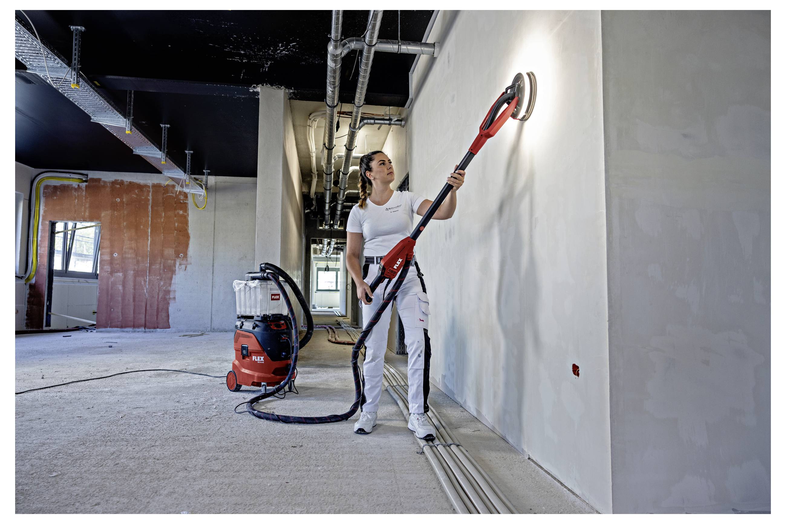 Un ouvrier du bâtiment ponçe un mur avec une ponceuse électrique à long manche dans une pièce inachevée, avec une unité d'aspiration à proximité et des tuyaux de plafond apparents.