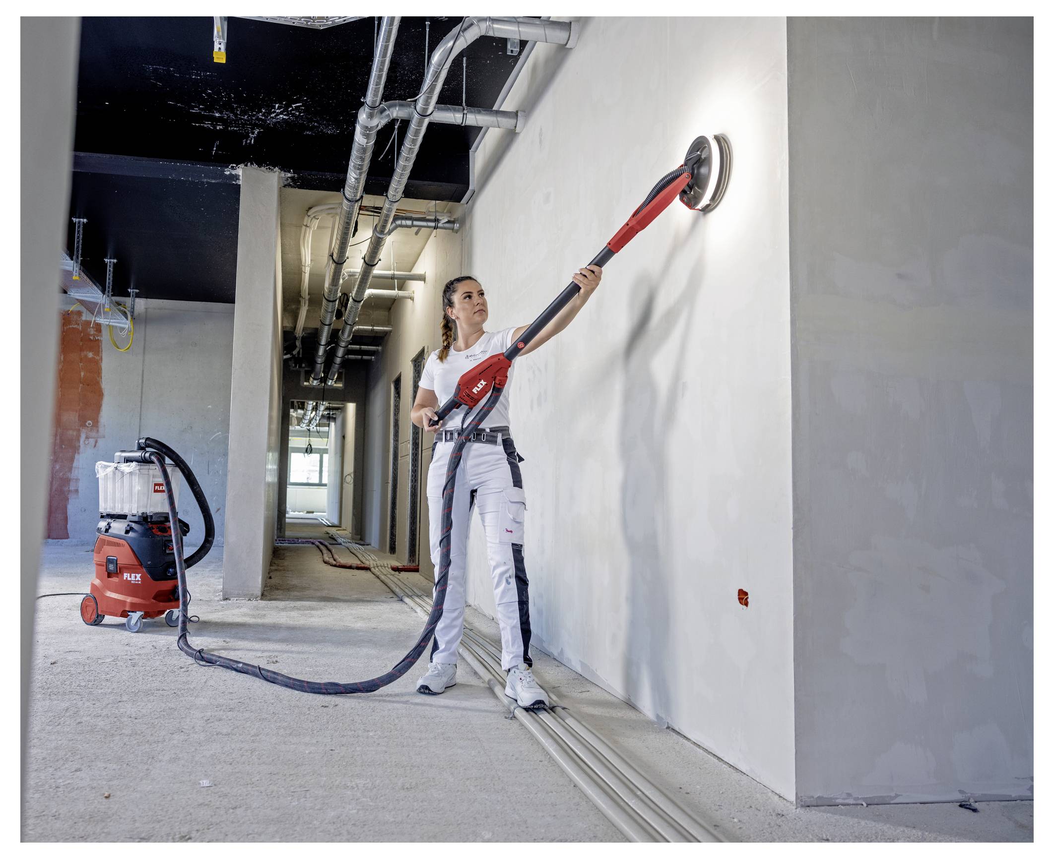 Ouvrier utilisant une ponceuse murale à long manche pour lisser un mur plâtré dans un couloir intérieur inachevé, avec un groupe de dépoussiérage sur le sol.
