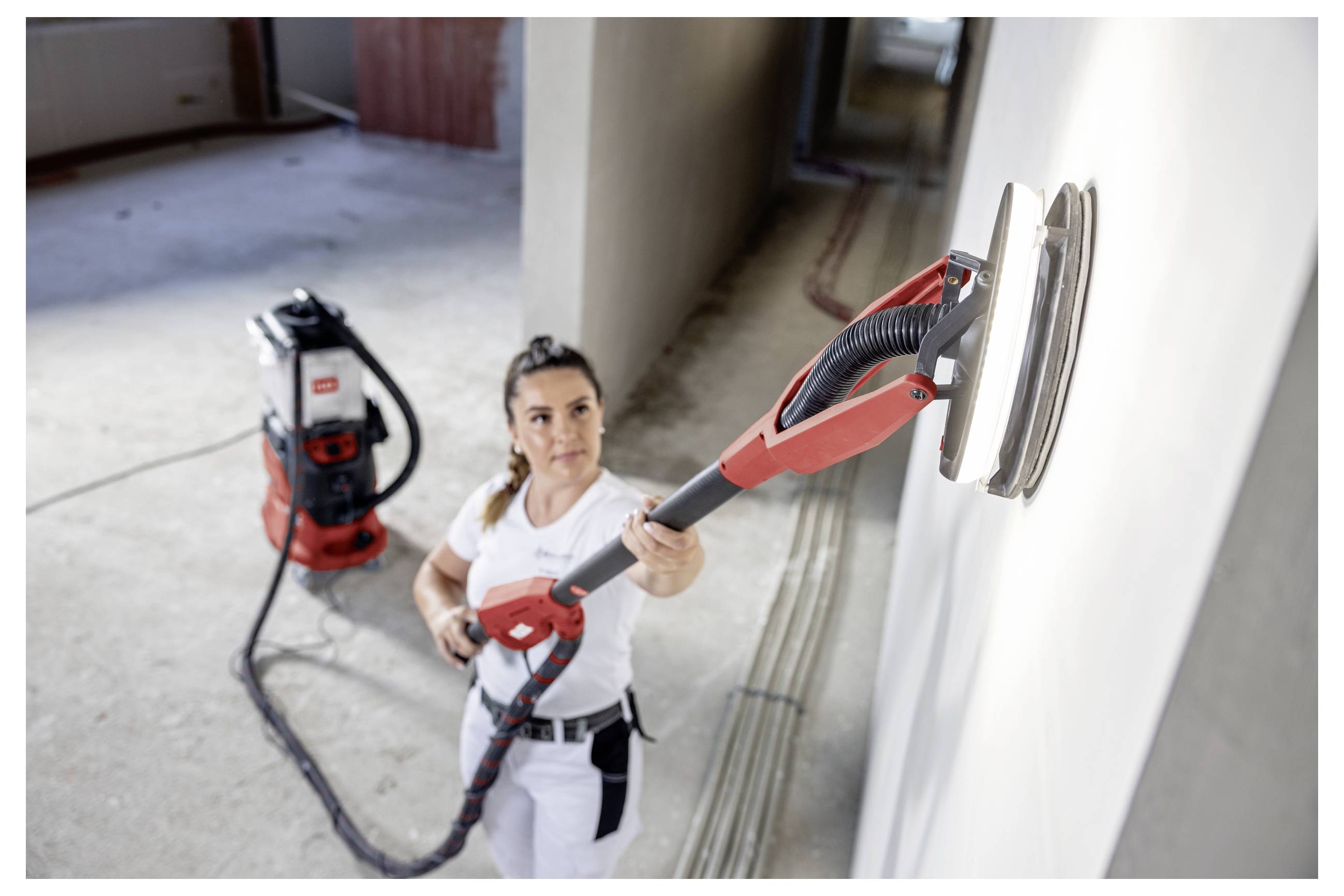 Femme utilisant une ponceuse murale rouge pour lisser un mur intérieur lors d'une rénovation domiciliaire, avec un extracteur de poussière à vide en arrière-plan.