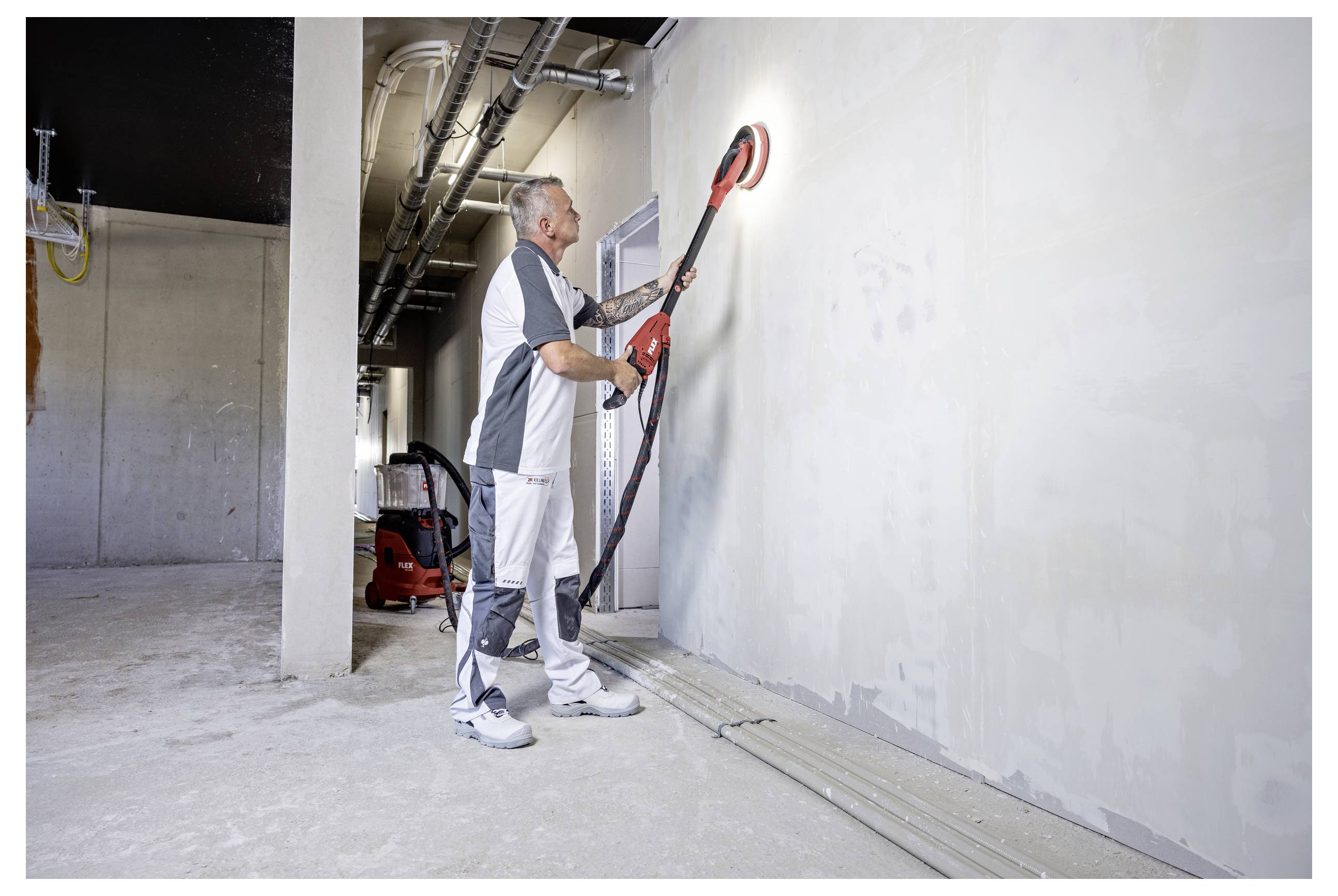 Un travailleur utilise une ponceuse électrique à long manche sur un mur à l'intérieur d'un bâtiment inachevé, lissant la surface près des tuyaux de plafond exposés.