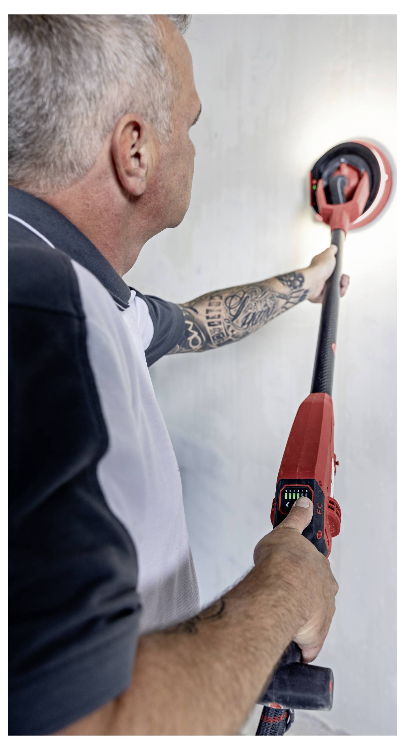 Un homme ponce un mur avec un ponceuse murale à perche rouge pendant une rénovation intérieure, lissant une surface réparée.