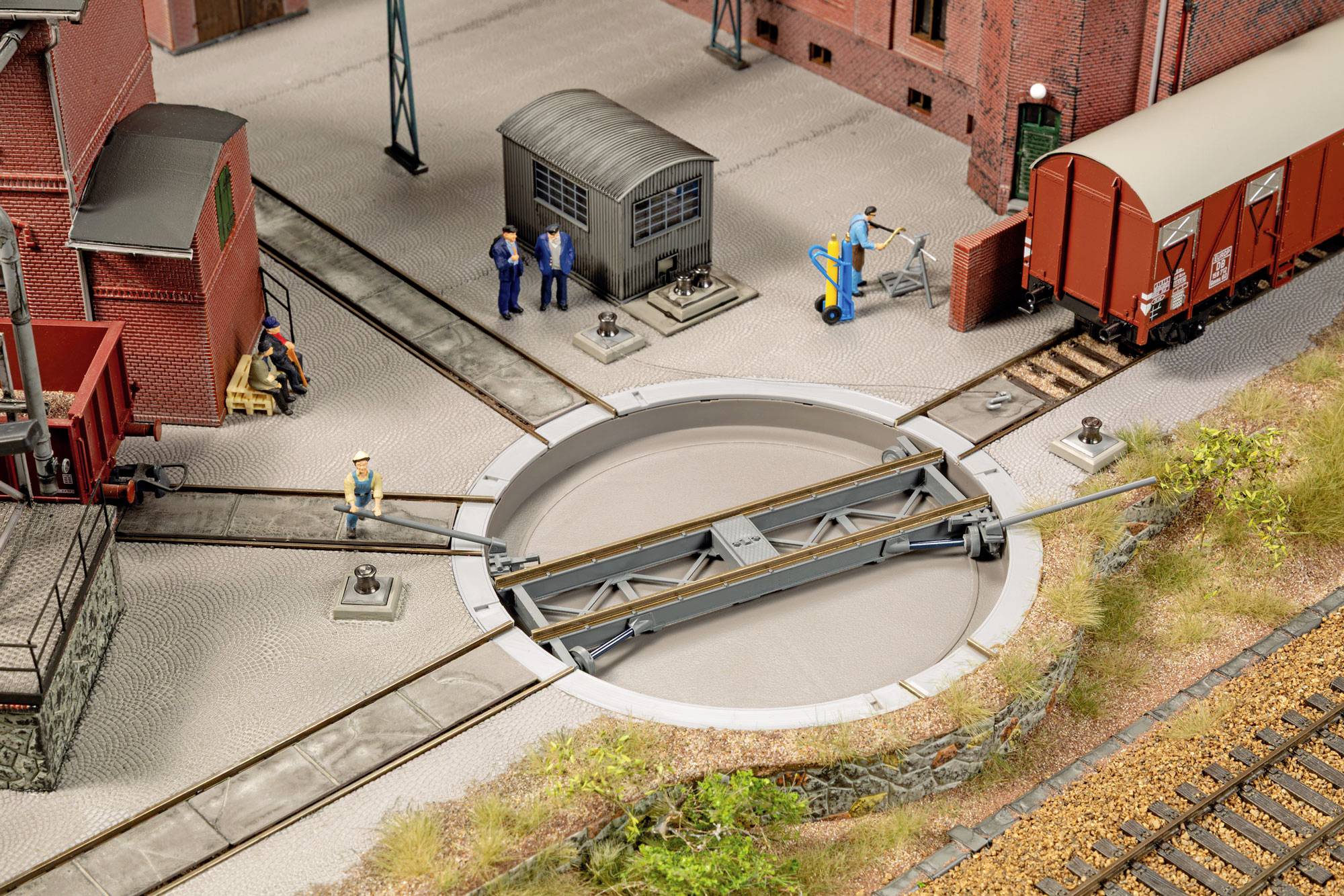 Cour de triage ferroviaire miniature avec un pont tournant central, des ouvriers, des remises et un wagon de marchandises rouge à côté de plusieurs voies.