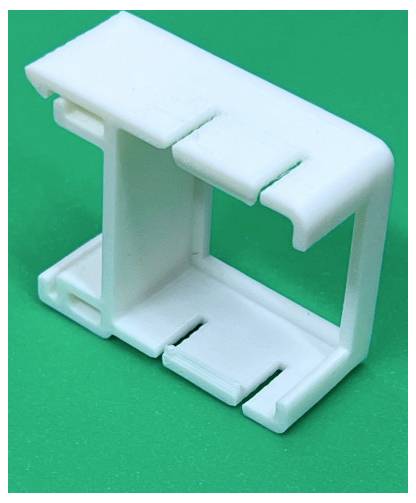 Clip ou support en plastique blanc avec un cadre rectangulaire ouvert, présenté sur un fond vert.