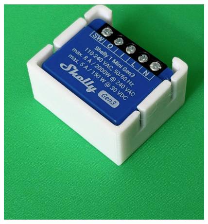 Module de relais intelligent Shelly Mini Gen3 dans un cadre de montage blanc sur un fond vert, avec des bornes à vis étiquetées SW, O, I, L et N.