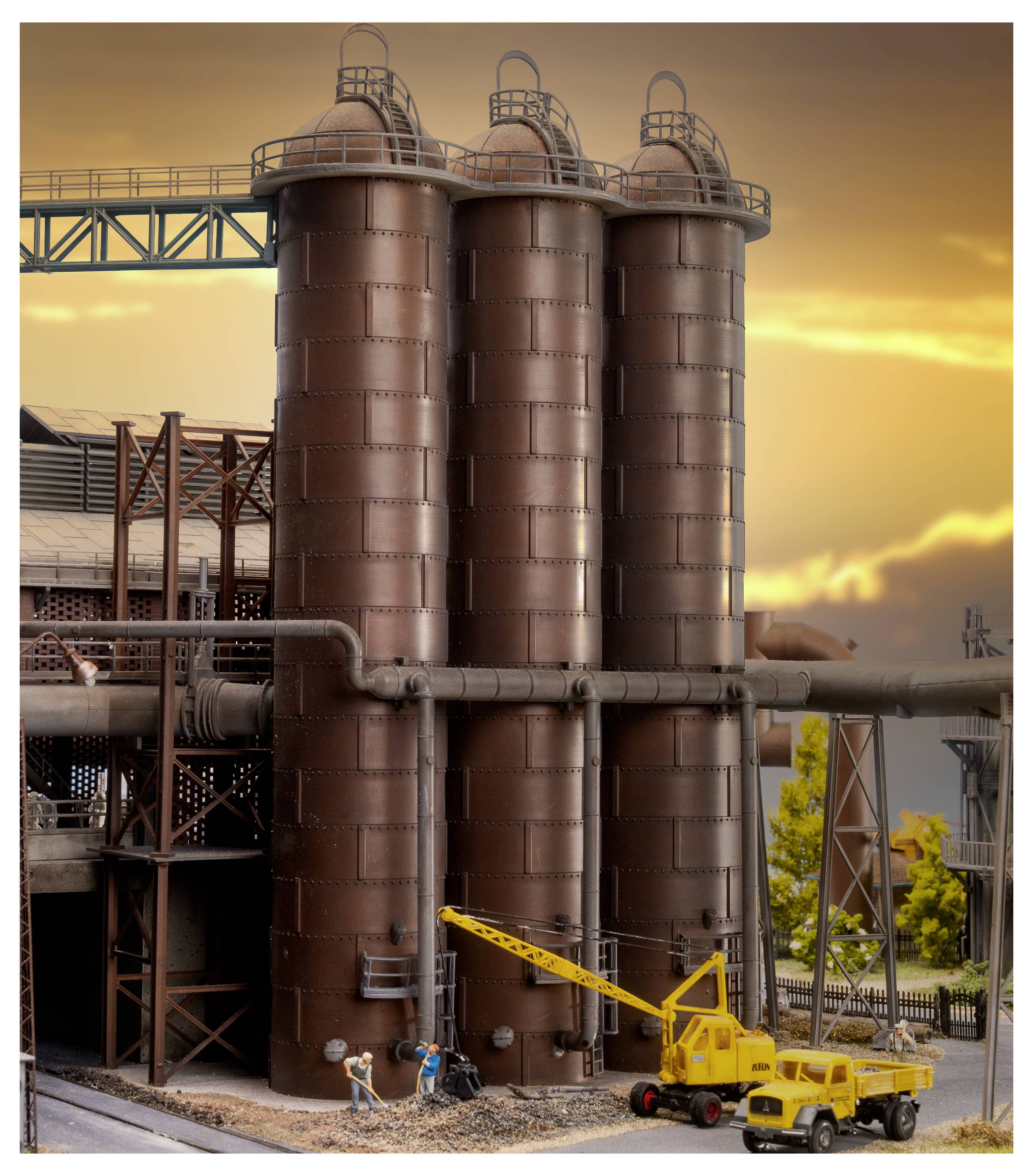 Usine industrielle avec trois hauts silos de stockage bruns, des travailleurs et un camion-grue jaune au coucher du soleil.