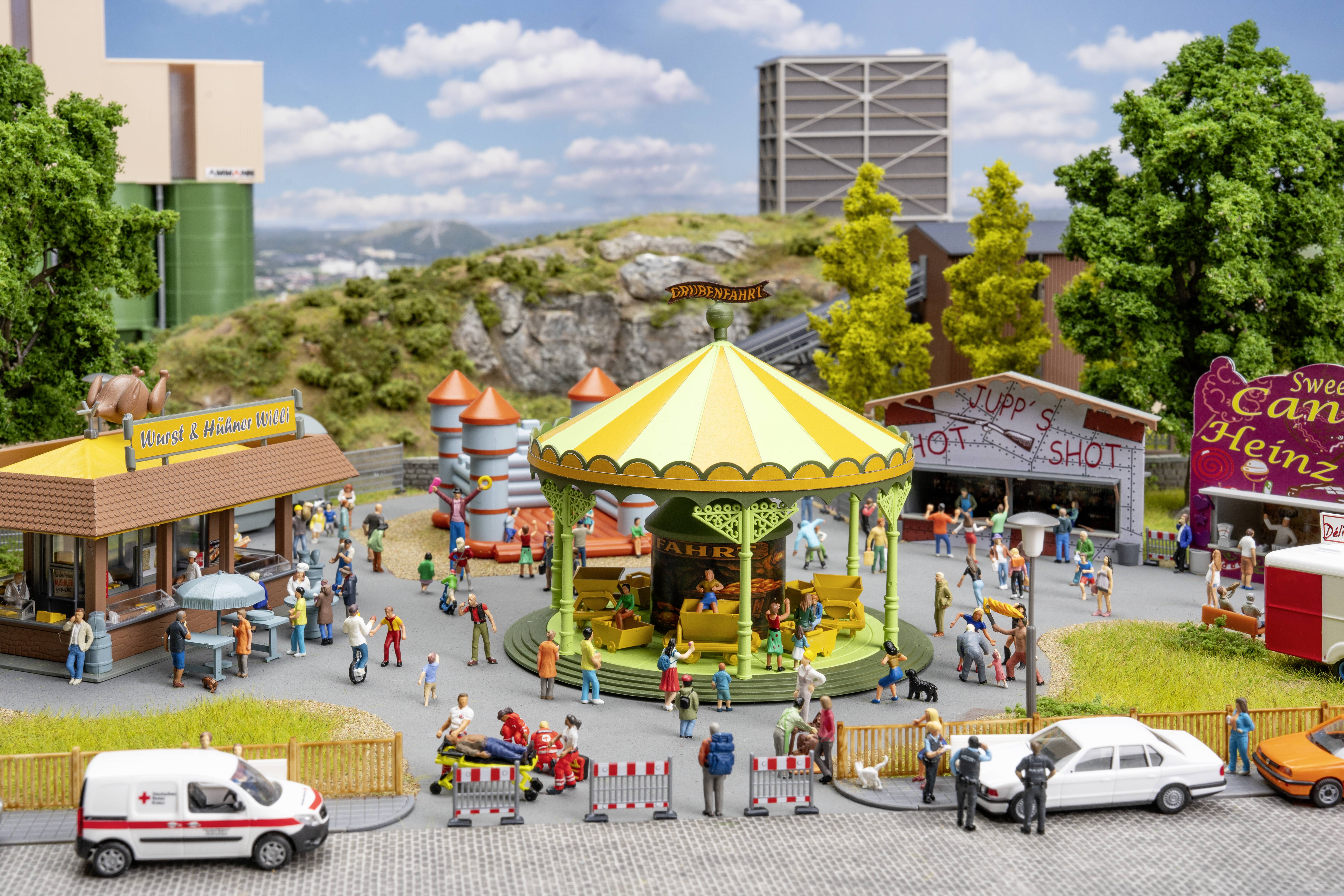 Scène de parc d'attractions miniature avec un carrousel rayé au centre, de petites foules, des stands de nourriture, un jeu de tir et des voitures garées au premier plan.