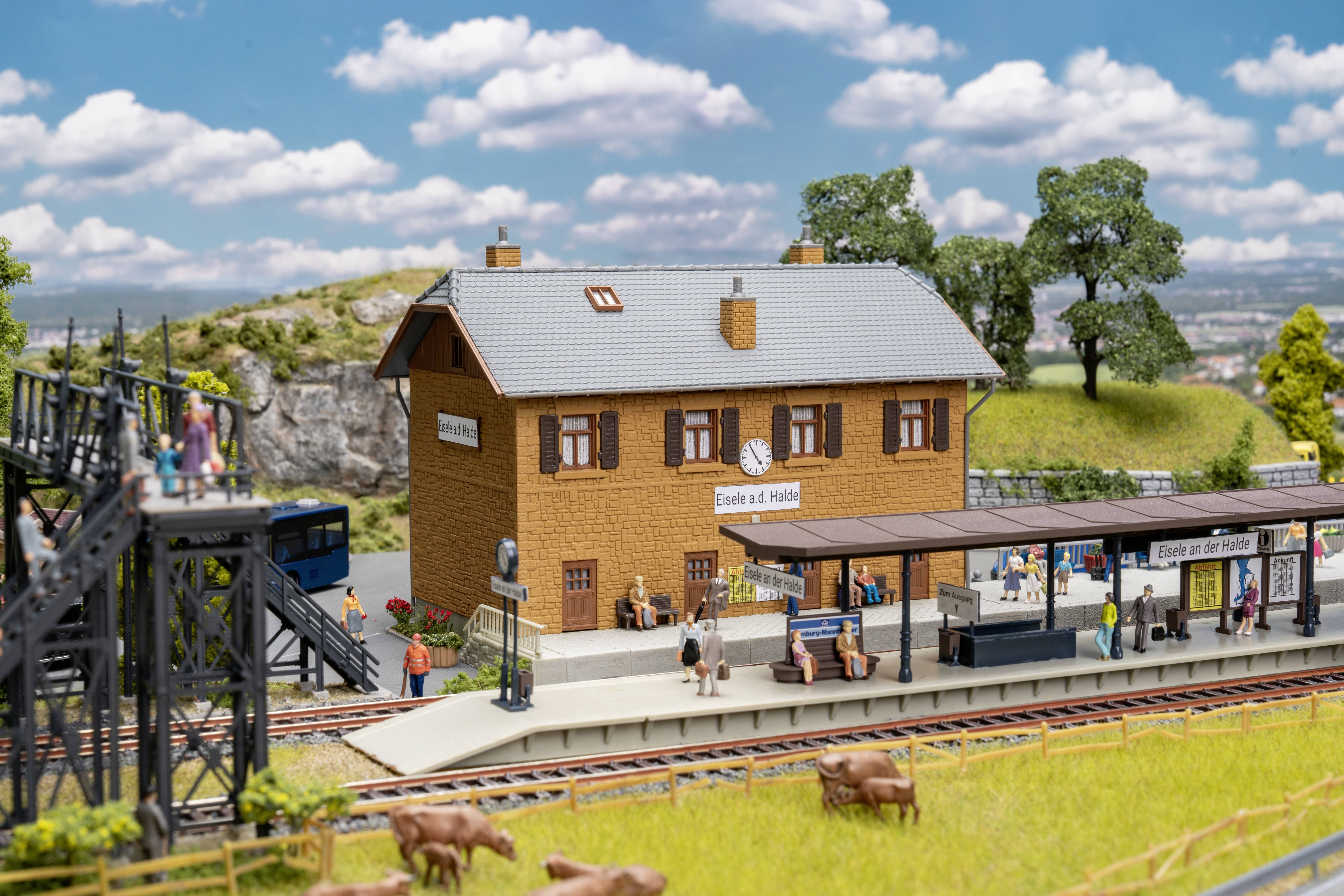 Scène de chemin de fer miniature d'un bâtiment de gare rurale portant l'étiquette « Tale o' the Halls », avec quai, voies, pont, personnages miniatures et vaches au premier plan.