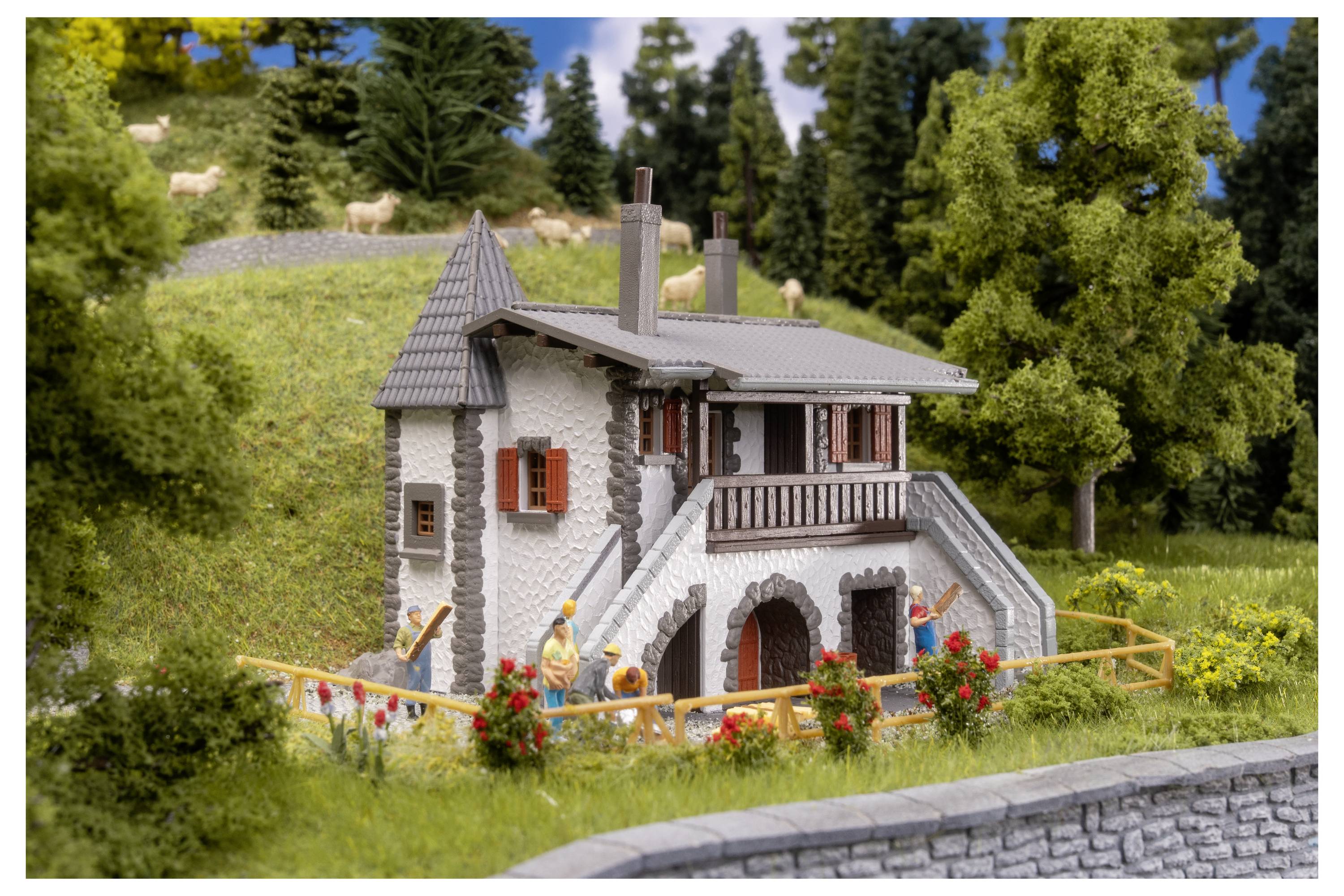 Maquette miniature d'une maison en pierre avec un escalier et un balcon, entourée d'herbe, de fleurs, d'arbres et de petites figurines dans une scène extérieure aménagée.