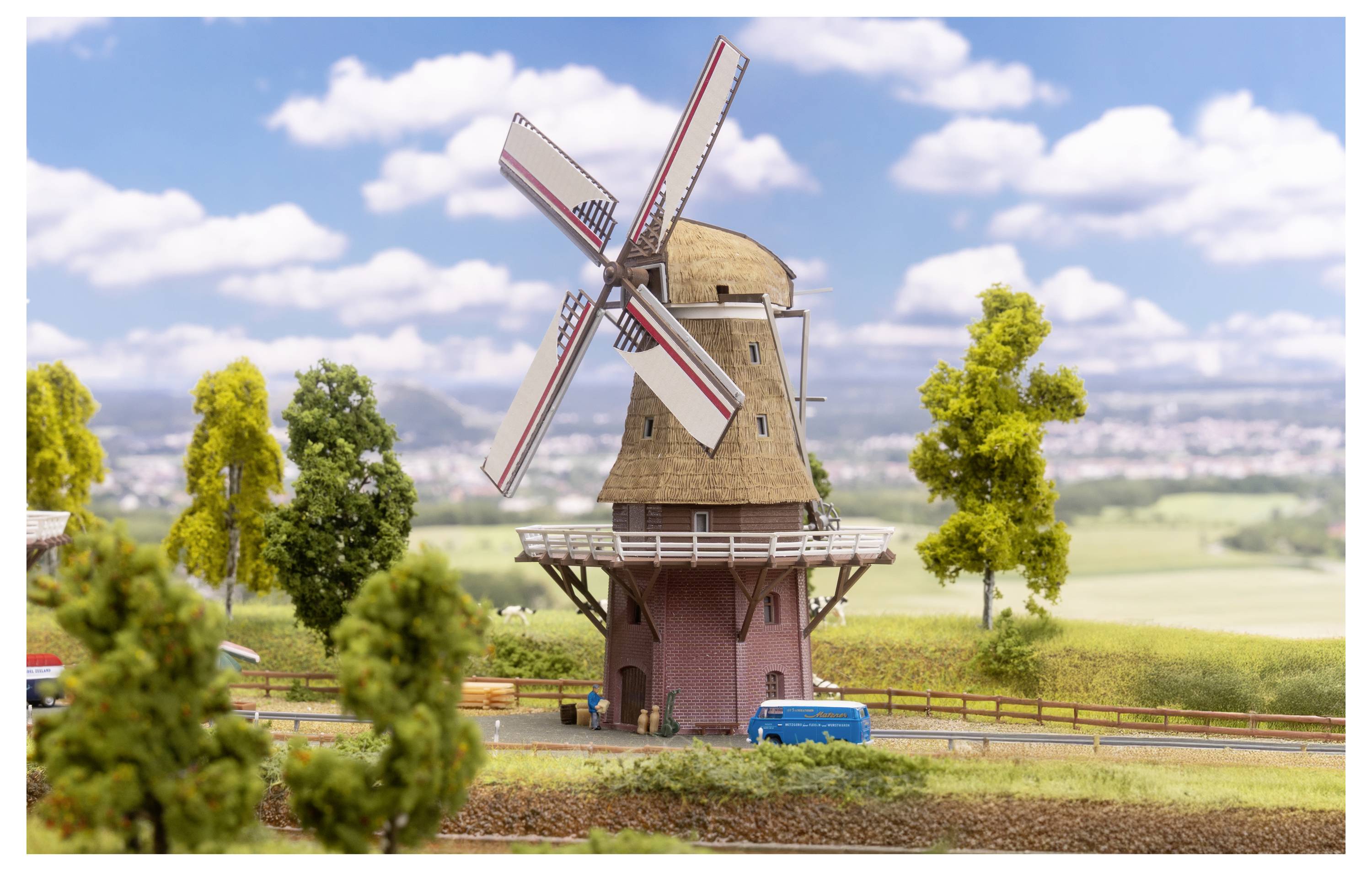 Moulin à vent miniature sur un paysage de campagne, avec de grandes pales, des arbres, des champs et un ciel bleu avec des nuages.