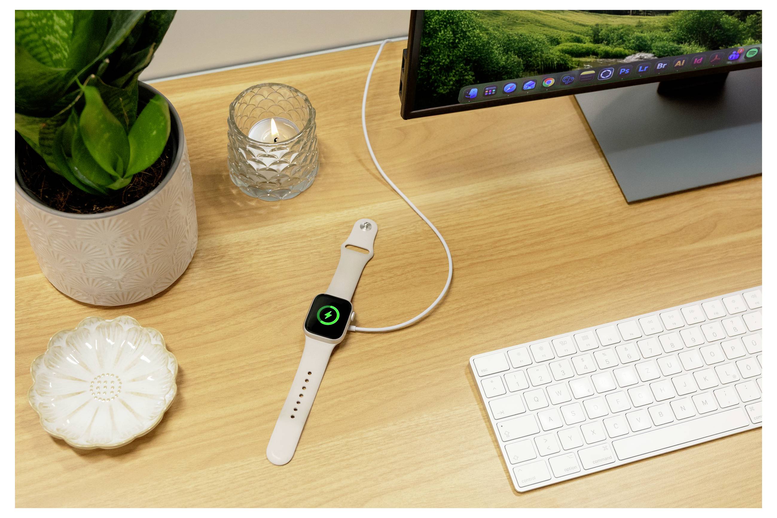 Apple Watch avec un bracelet blanc en charge sur un bureau en bois à côté d'un clavier, d'un moniteur, d'une plante et de bougies.