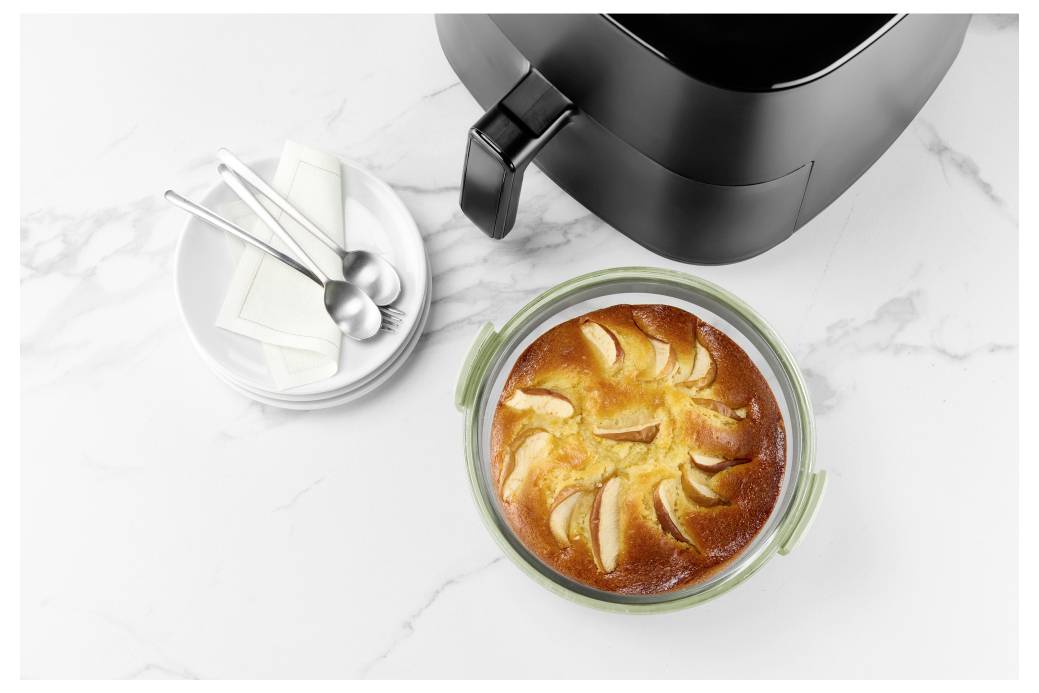 Vue de dessus d'une friteuse à air avec un dessert de pomme cuite dans un plat en verre à côté de petits bols d'épices et d'ustensiles sur un plan de travail blanc.