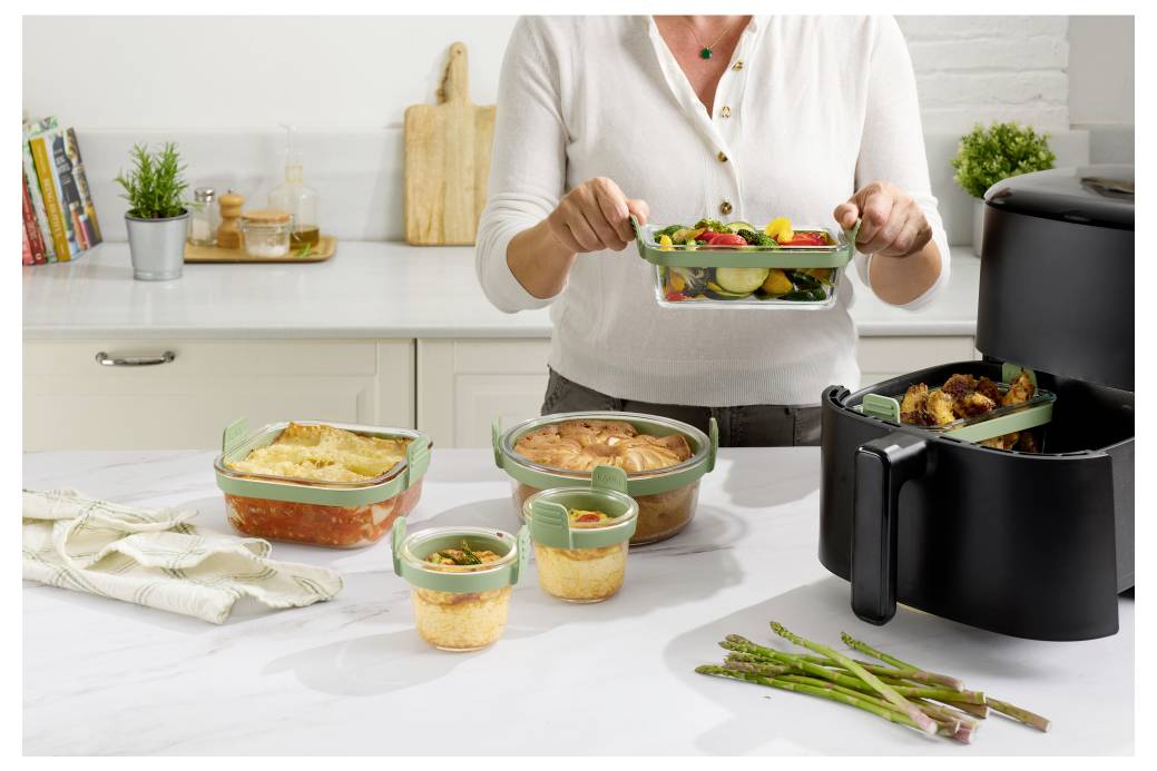 Une personne arrange de la nourriture préparée à l'avance sur un plan de travail de cuisine à côté d'un four à air ouvert, avec plusieurs récipients en verre étiquetés contenant des plats cuits et des légumes.