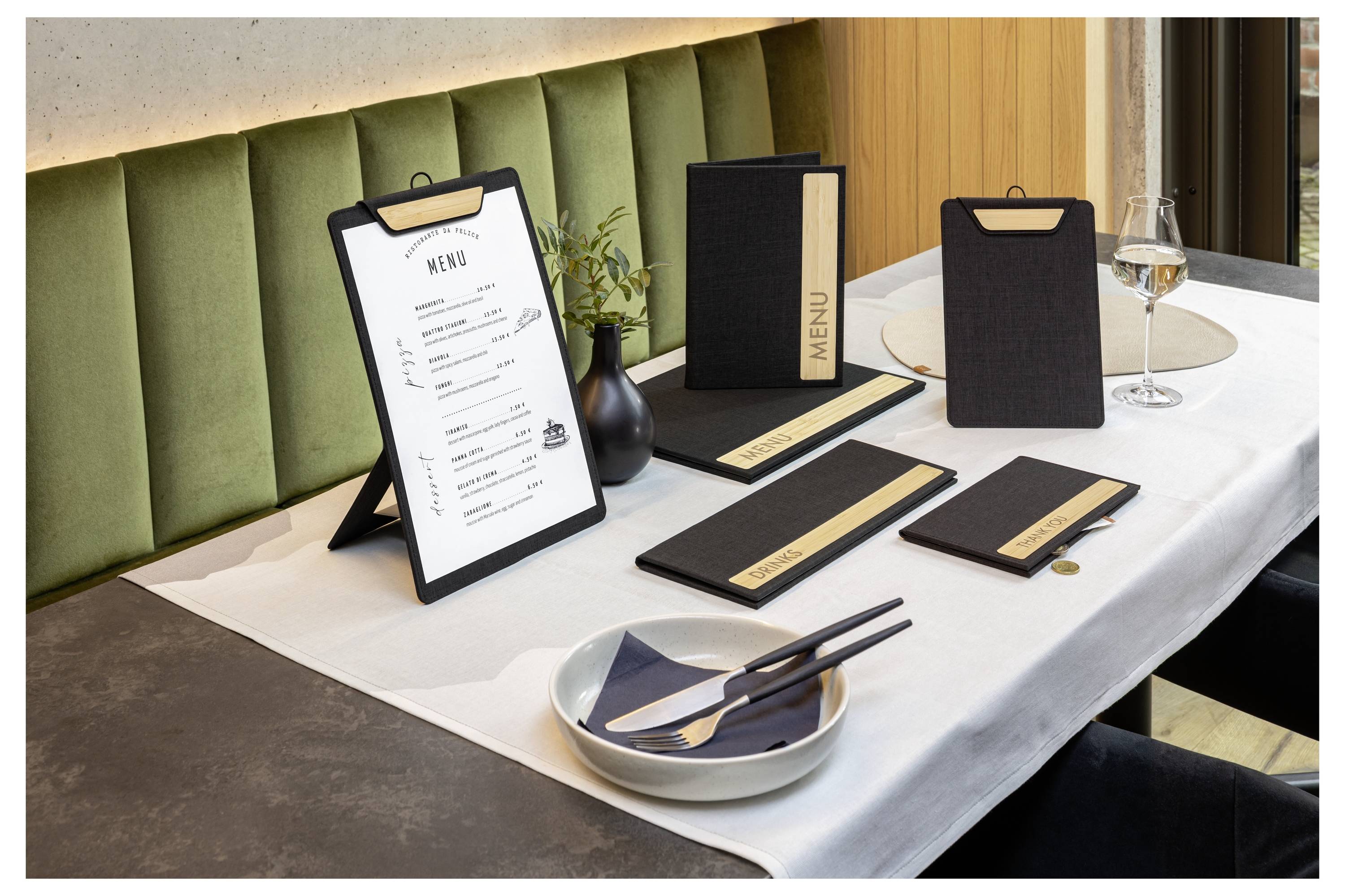 Restaurant table setting with framed menu, black menu holders, cutlery, plate, and wine glass on a white tablecloth.