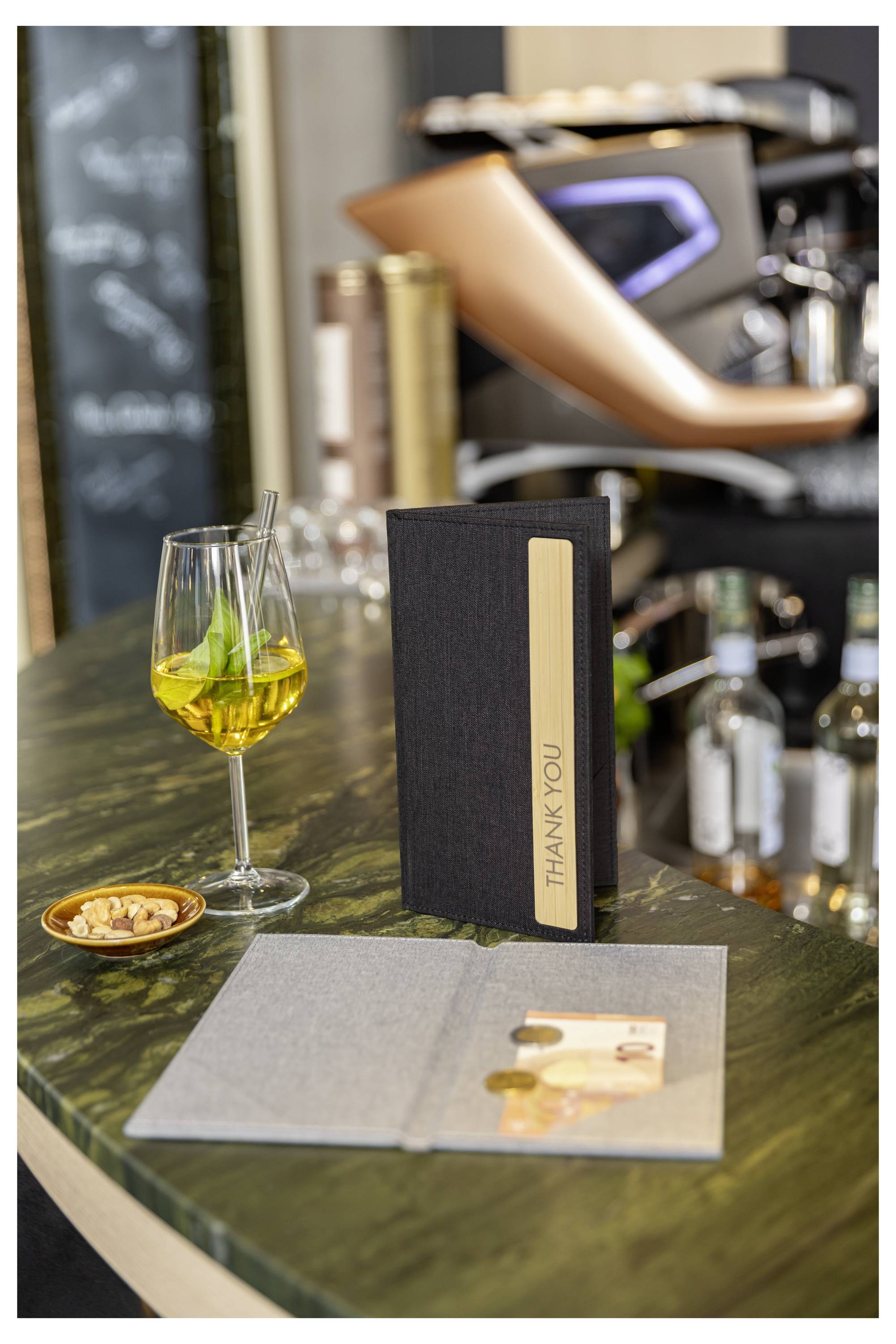 Restaurant table setting with a drink and a black 'THANK YOU' menu card, with an open menu or flyer on the table.