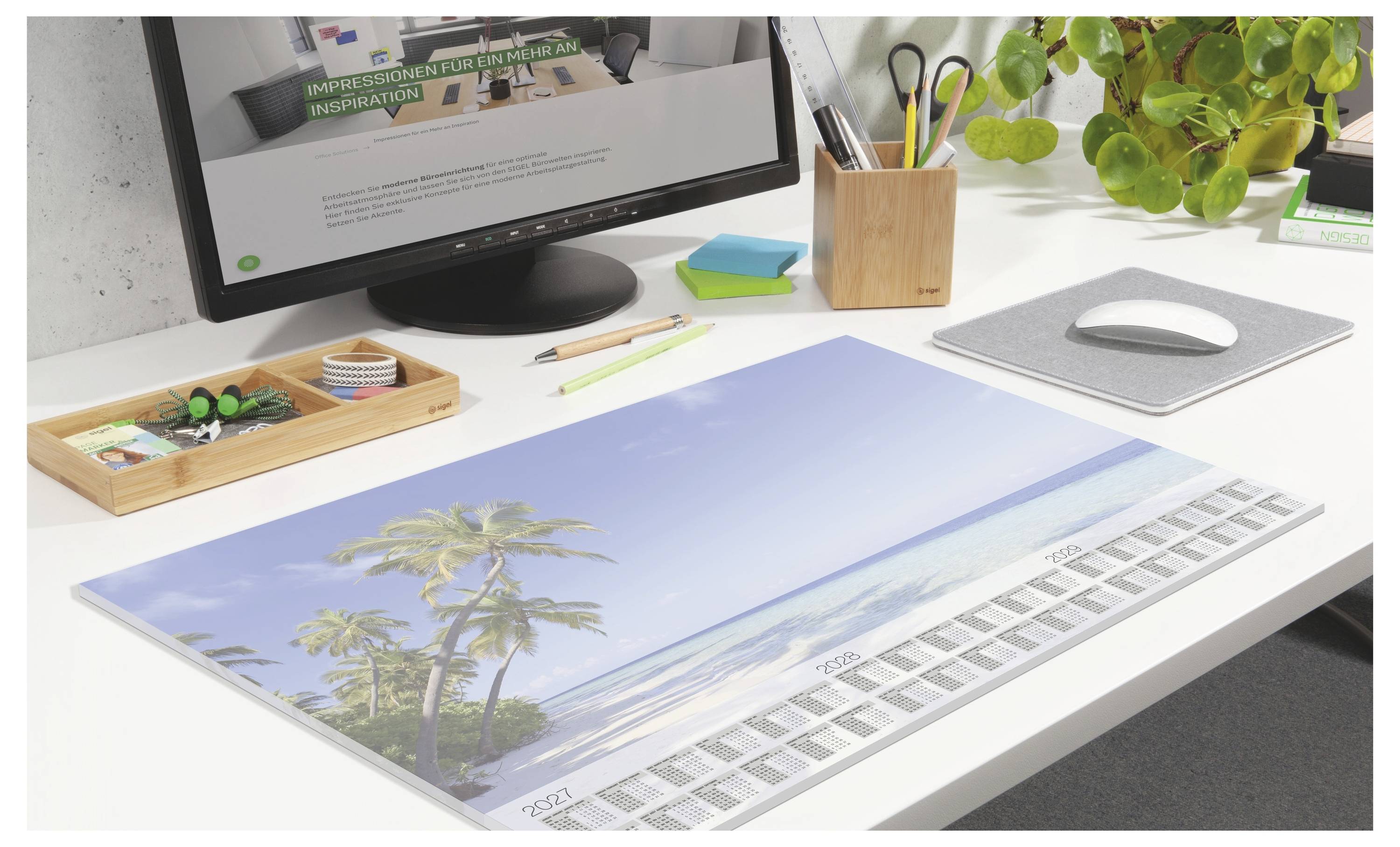 A desk workspace with a large calendar or planner featuring a tropical beach photo, plus pens, stationery, a laptop, and a plant.