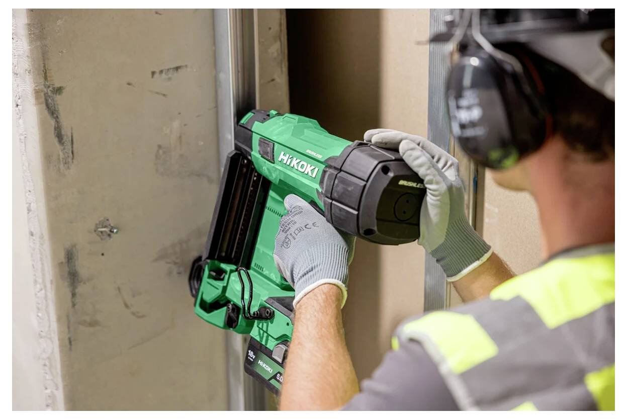 Construction worker using a green HiKOKI nail gun to fasten a door frame in a building interior, wearing gloves and safety headset.