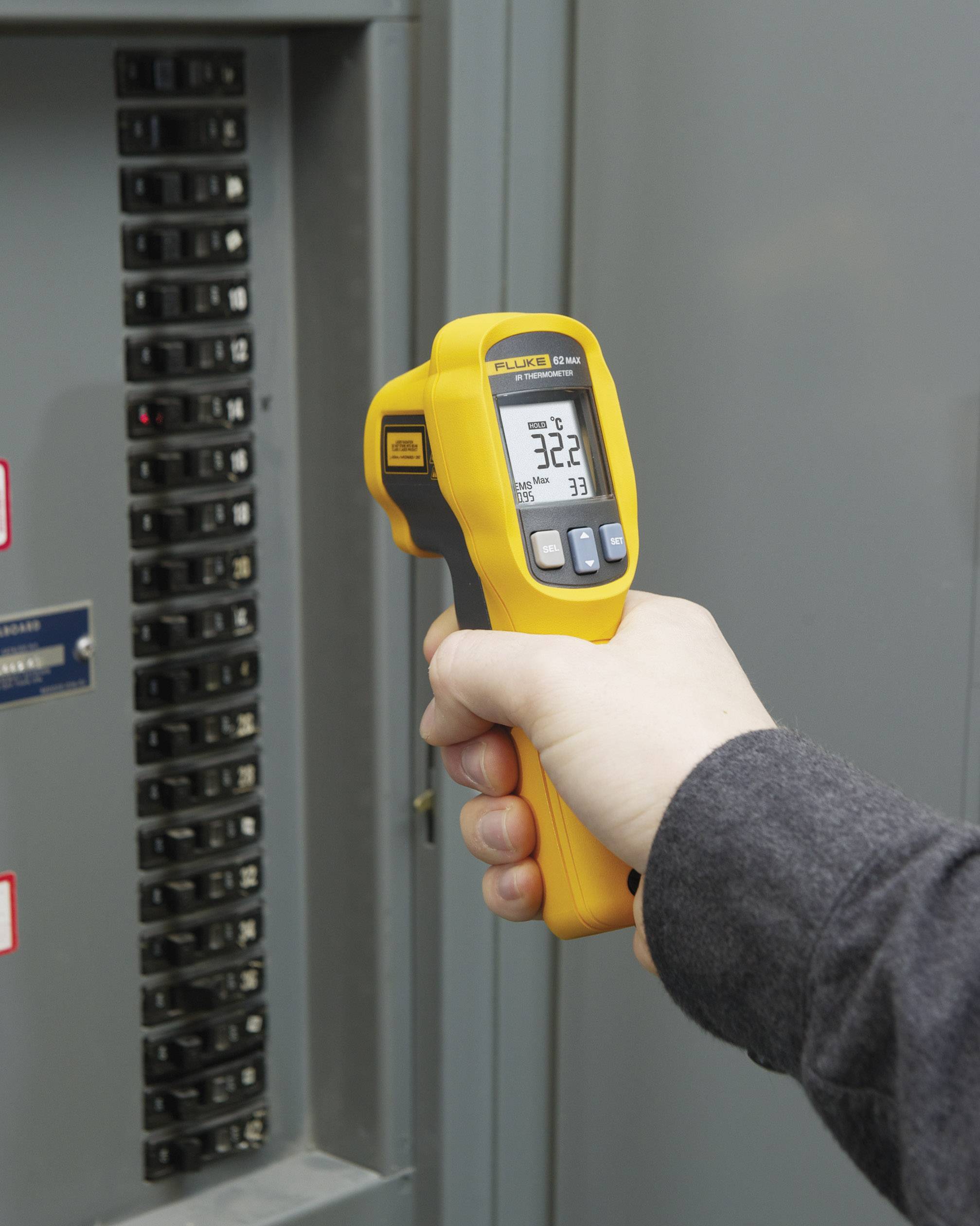 Une main tient un thermomètre infrarouge jaune devant un coffret électrique et affiche 32,2 degrés Celsius sur l'écran.