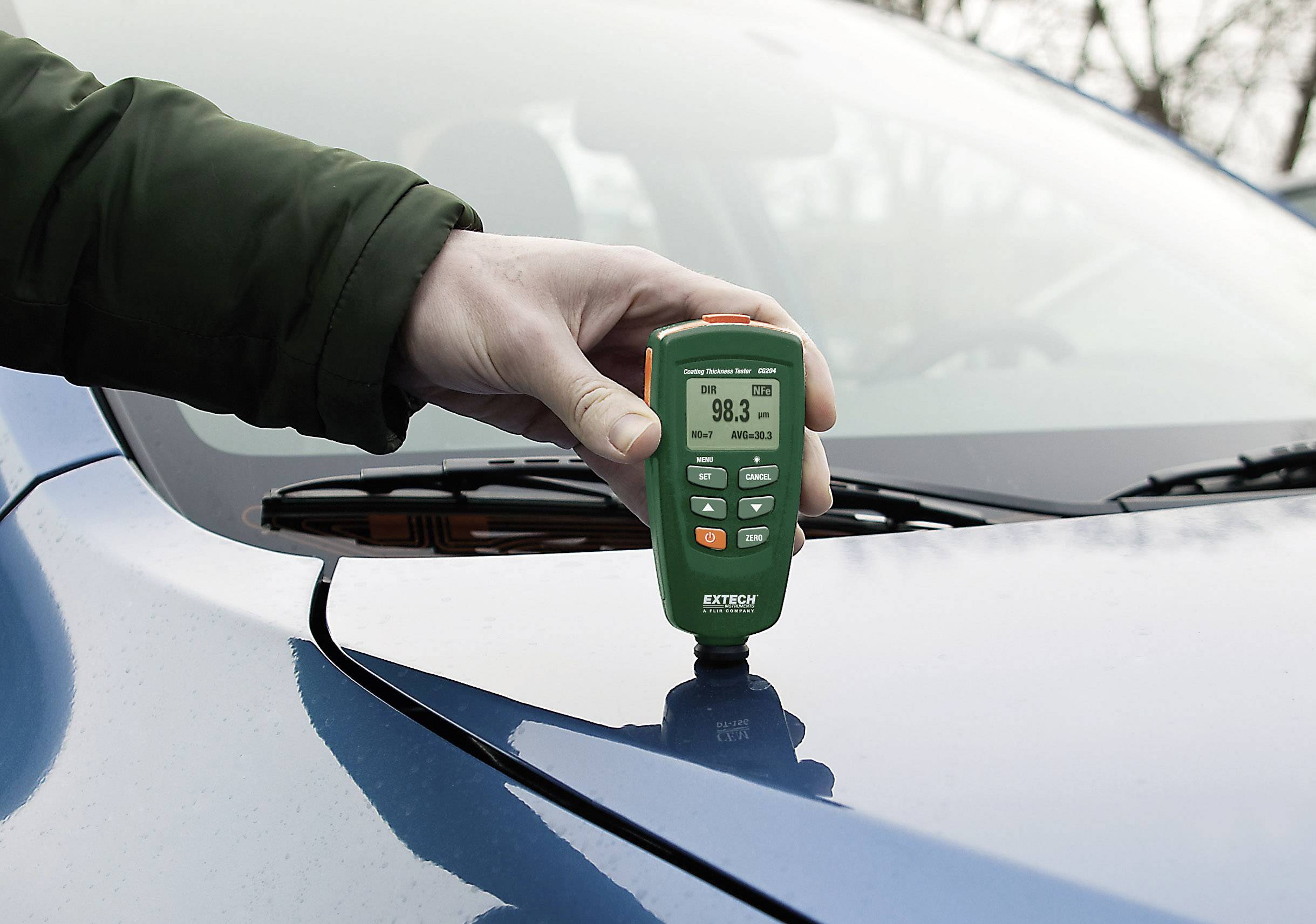 Une main tient un appareil de mesure d'épaisseur sur un toit de voiture bleu, affichant une épaisseur de 98,3 micromètres.