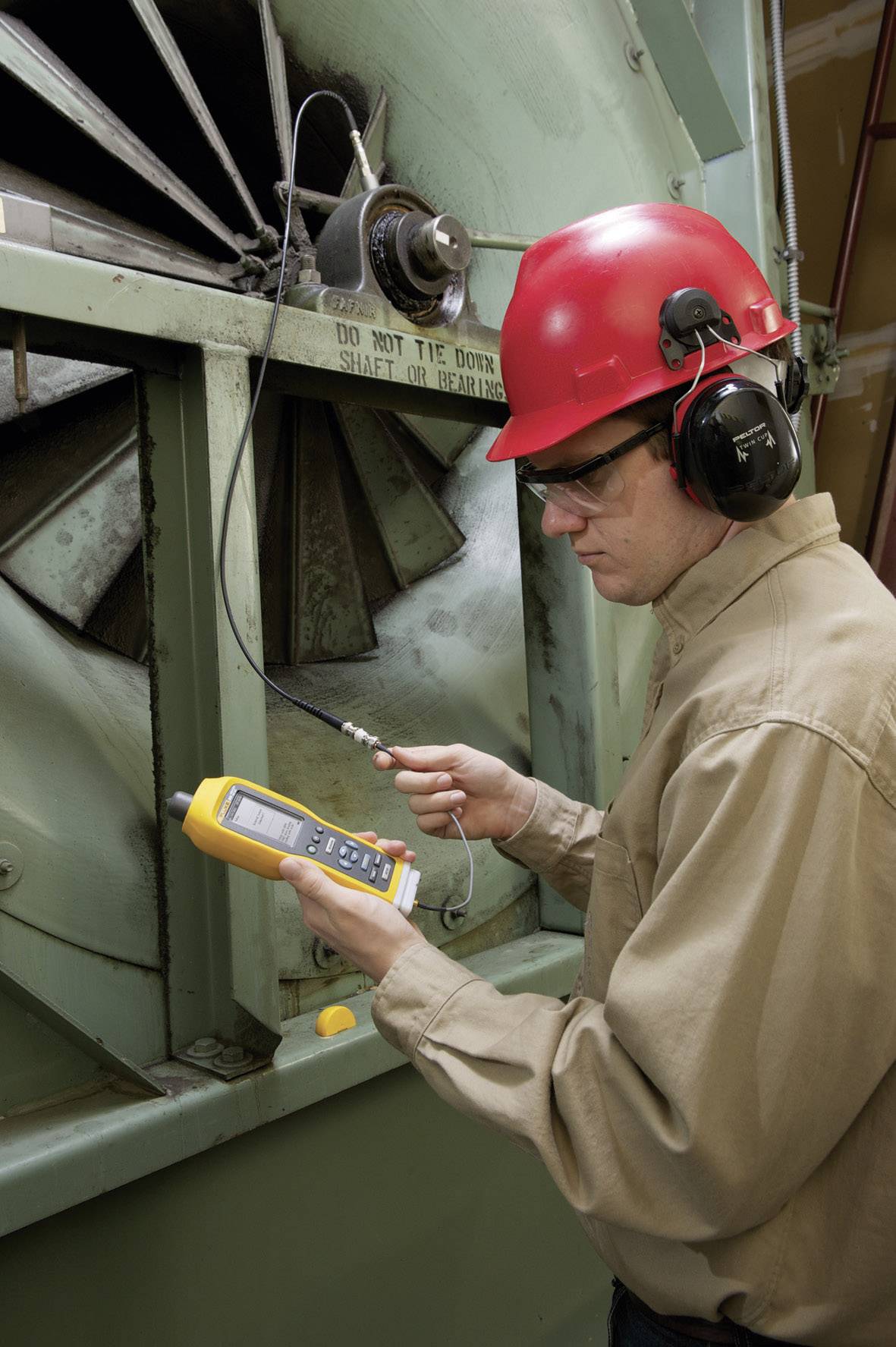 Un homme portant un casque de protection rouge et des protections auditives utilise un appareil de mesure portable pour relever des données à partir d'une grande unité de machine.