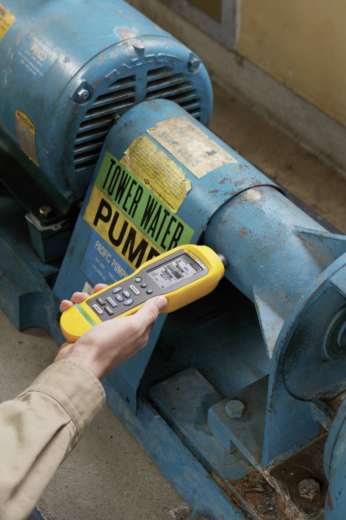 Une main tient un appareil de mesure jaune qui vérifie la vibration d'une machine industrielle bleue sur laquelle se trouve un panneau vert portant l'inscription 'PUMP'.