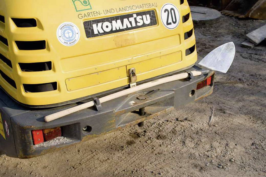 Un petit pelleteuse jaune portant l'inscription 'Komatsu' avec en arrière-plan 'Aménagement paysager'. Une pelle est posée à l'arrière.
