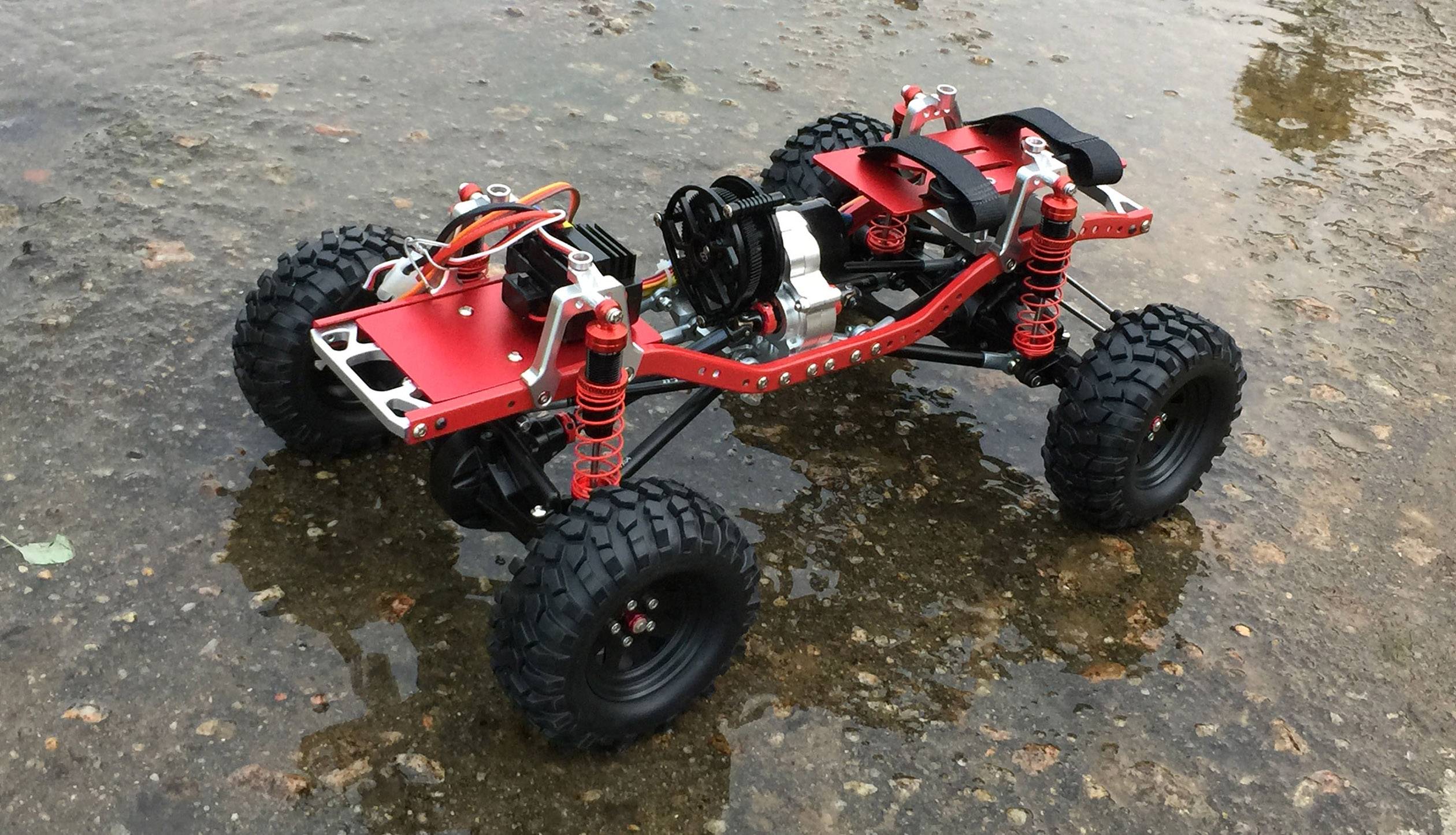 Une voiture télécommandée avec des roues robustes tout-terrain et un châssis rouge est posée sur un sol humide et pierreux.