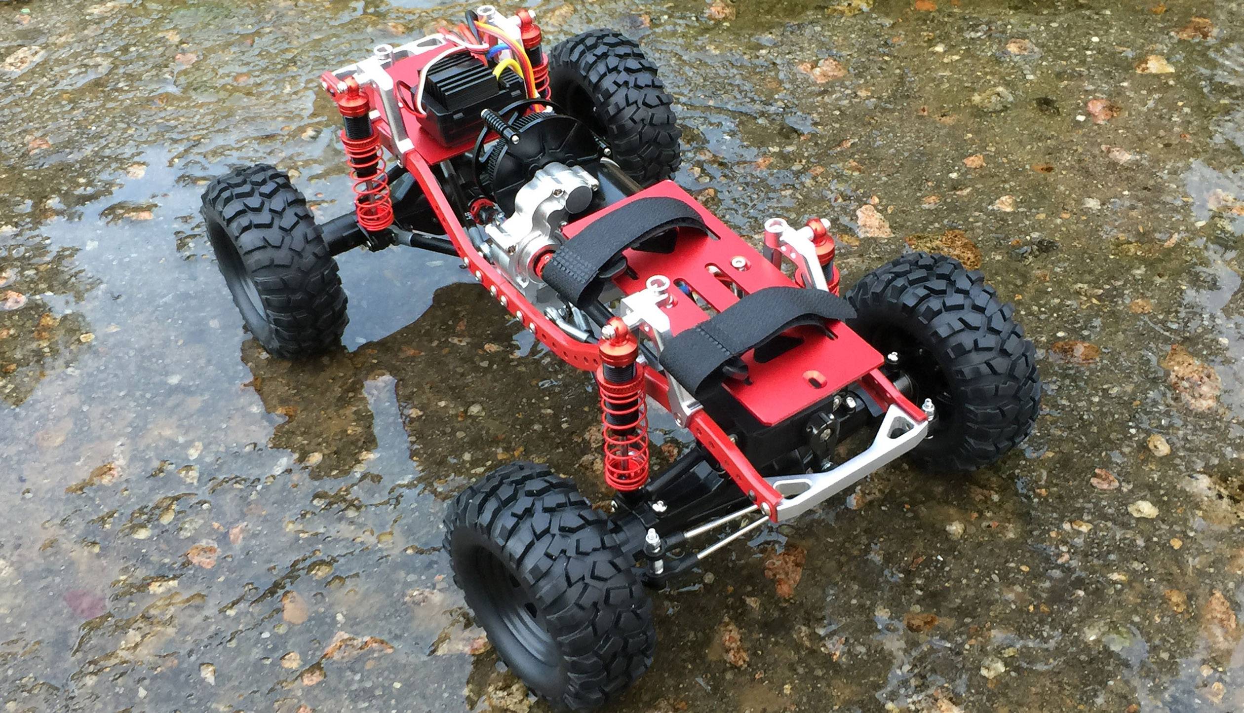 Voiture télécommandée avec châssis robuste et grandes roues, placée sur une surface humide et rocheuse. Adaptée aux terrains tout-terrain.