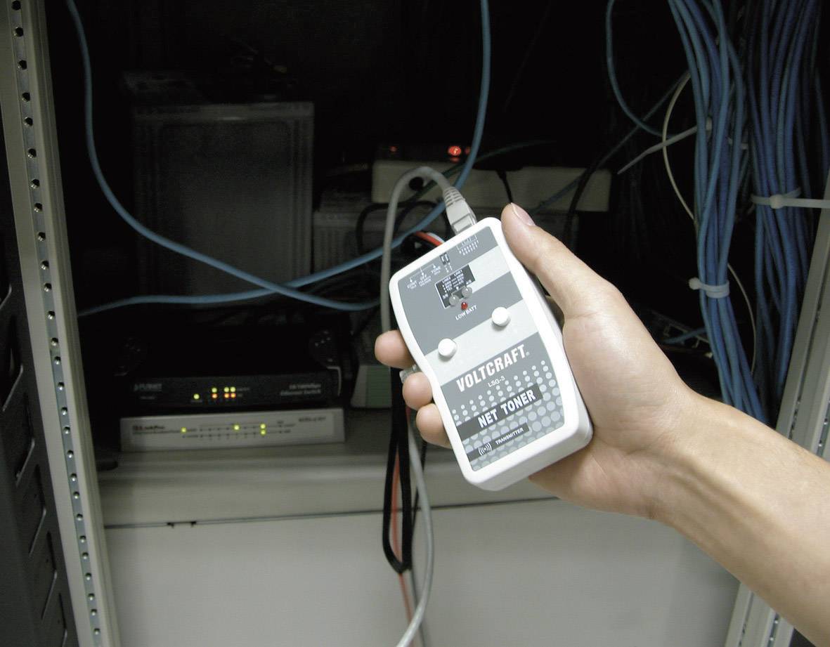 Une main tient un appareil de test de câbles réseau dans une salle de serveurs. À l'arrière-plan, divers équipements électroniques et câbles sont visibles.